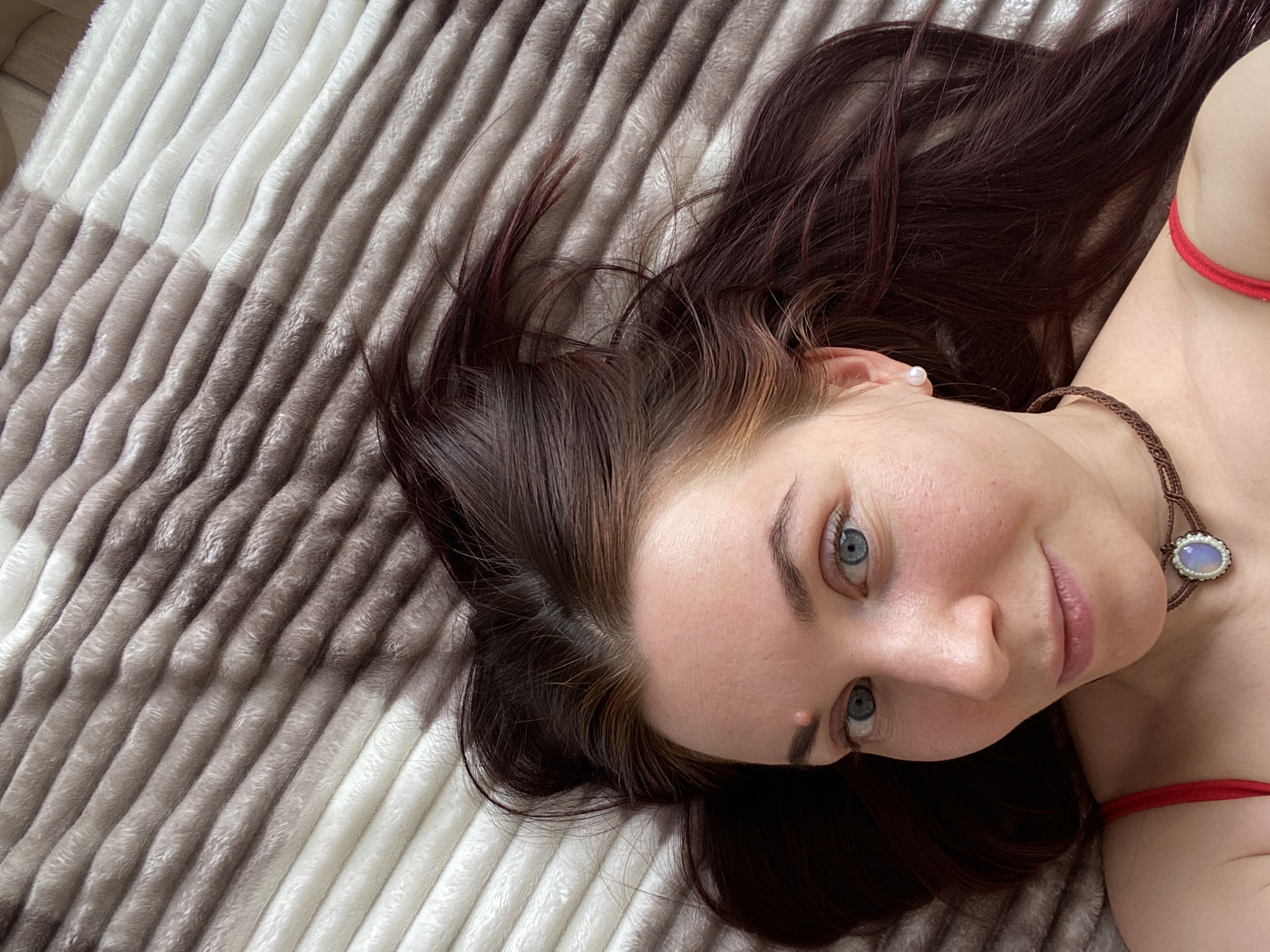 A young woman is lying on a horizontally striped beige and white blanket. She has dark auburn hair, blue eyes, and fair skin. She is wearing a pearl earring and a brown woven necklace with a pendant. A red strap is visible near her shoulder. The location cannot be determined based on the information provided.
