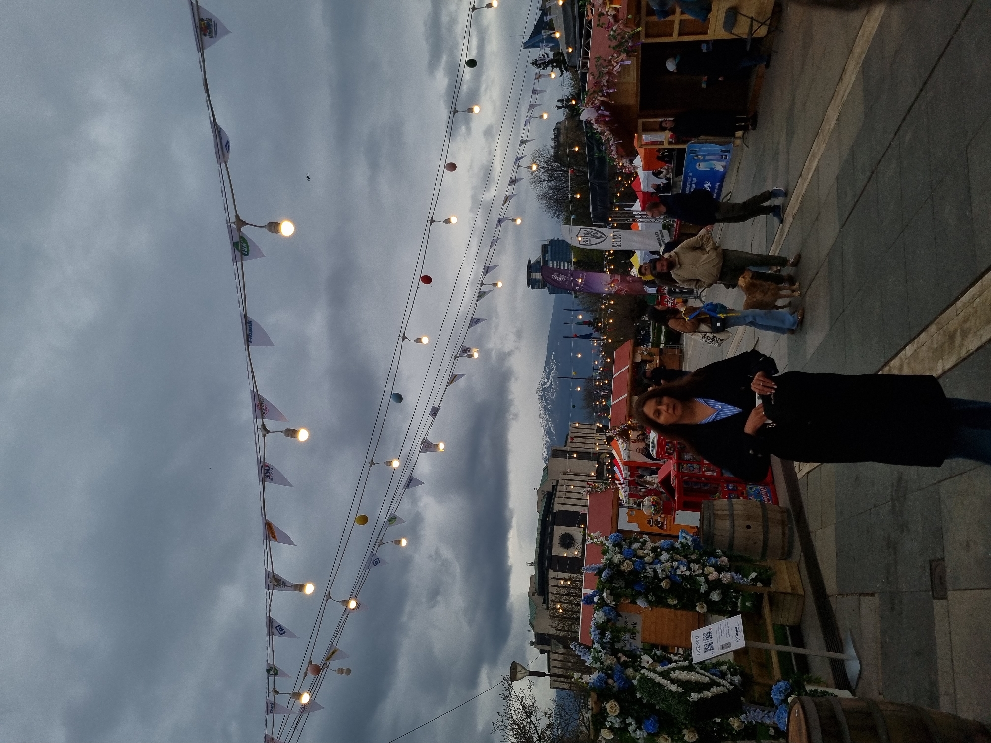 The image depicts a street scene in Sofia, Bulgaria, likely at a public market or festival. A woman in a dark coat stands in the foreground. Strings of lights with flags are strung overhead. There are people walking in the background, including a person with a dog. Stalls decorated with flowers and other items line the street. Buildings and mountains are visible in the distance. The sky is overcast and cloudy.