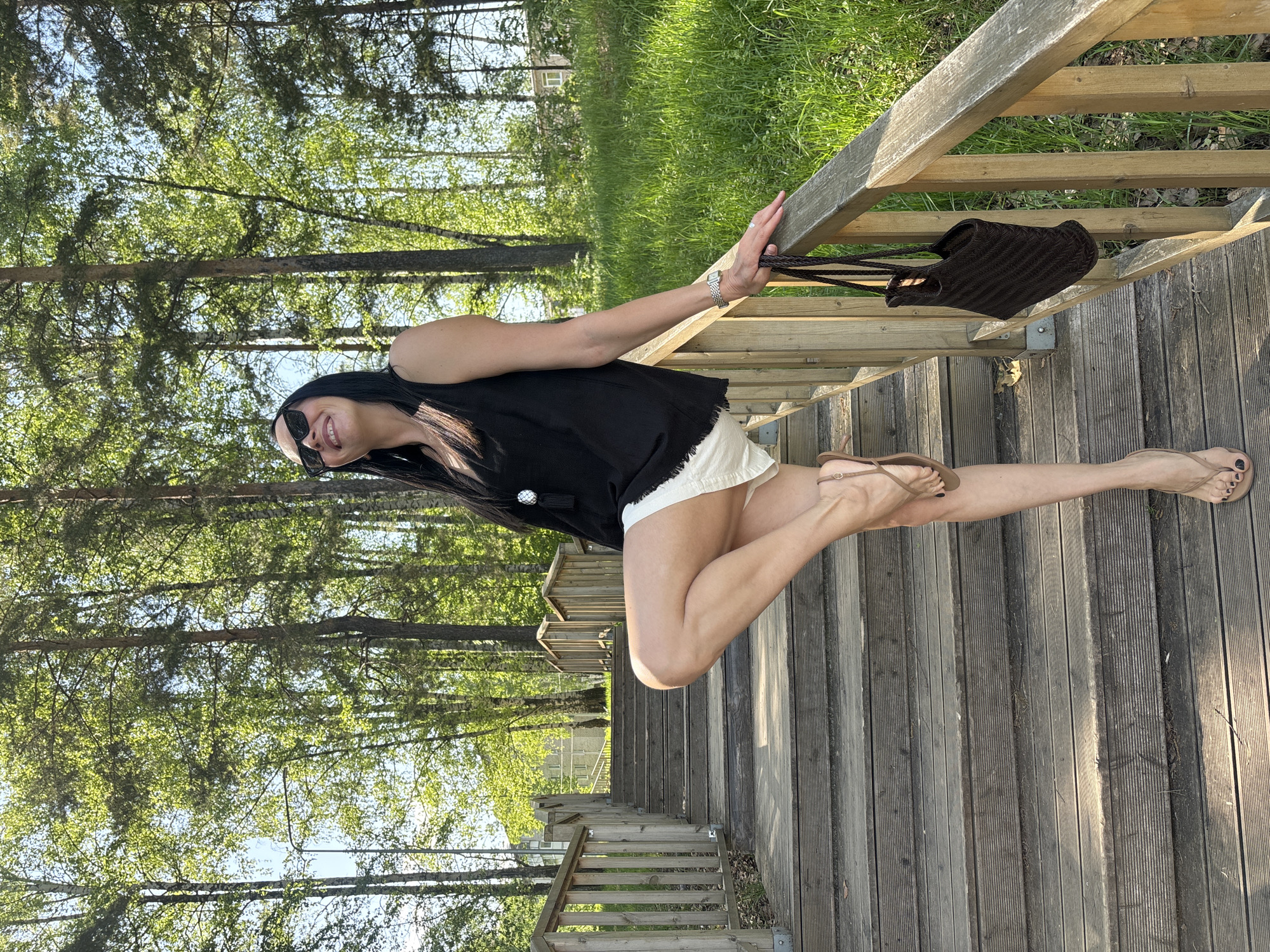 A woman stands on a wooden staircase in a park-like setting. She is wearing a black sleeveless top, white shorts, and brown sandals. She has black hair and is wearing sunglasses. She is holding onto the wooden railing of the staircase with her left hand, and a brown bag hangs from the railing next to her hand. The staircase leads downwards into a forest area with tall trees and green grass. The location is possibly in Chaikovsky, Russia.