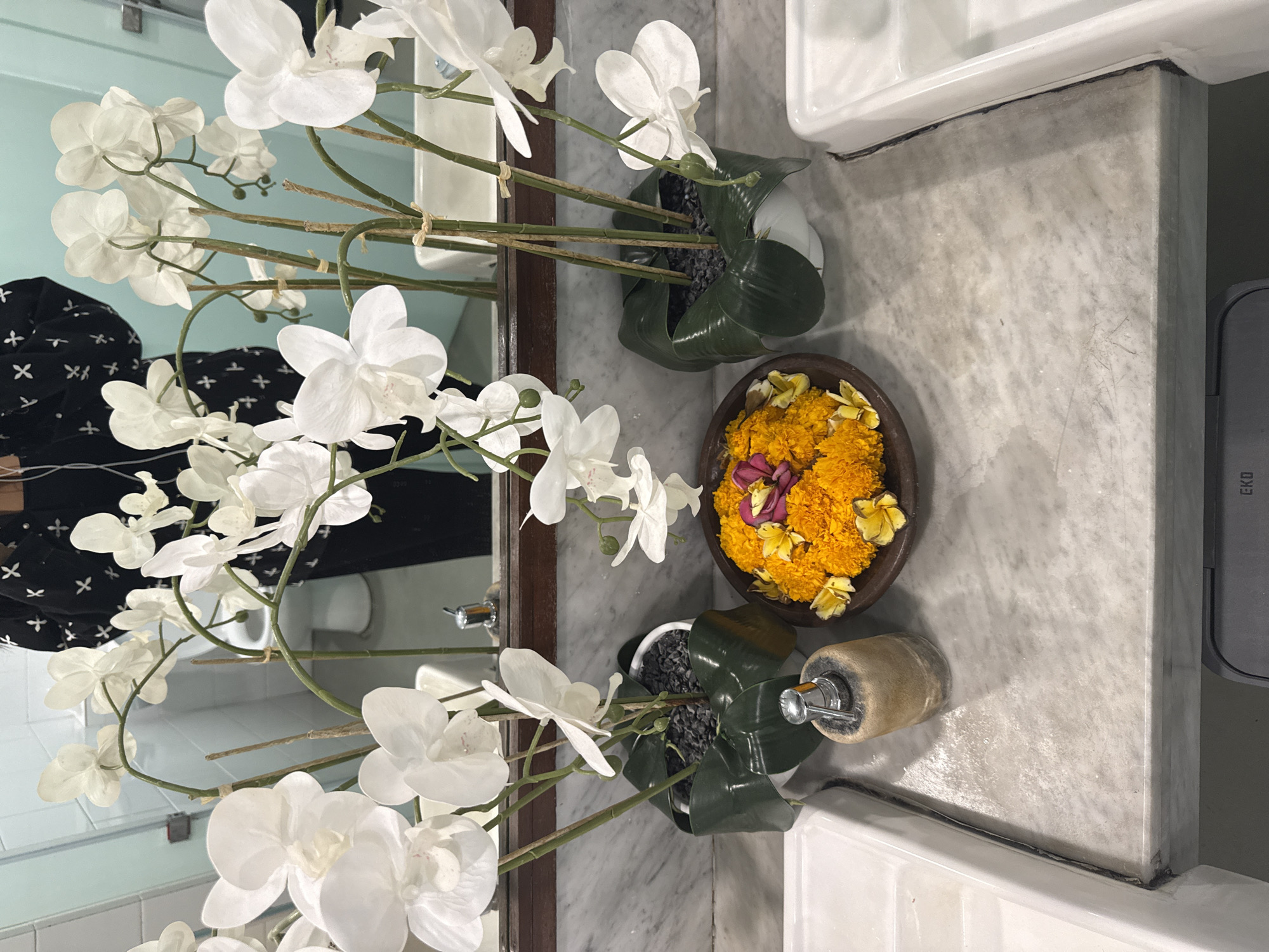A man stands in front of a mirror in a bathroom. He is wearing a black shirt with a white cross pattern and is reflected in the mirror. In the foreground, there are two white orchid plants in white pots, each adorned with dark green leaves. In the center, a rustic bowl overflows with bright orange marigolds and a few yellow flowers, with a pink flower at its center. To the left of the flower bowl is a dispenser with a silver pump and a beige stone-like base. The countertop appears to be made of white marble, and there are two white sinks visible on either side of the countertop. The bathroom walls are tiled in a light blue color. The overall scene suggests a serene and decorated bathroom space.