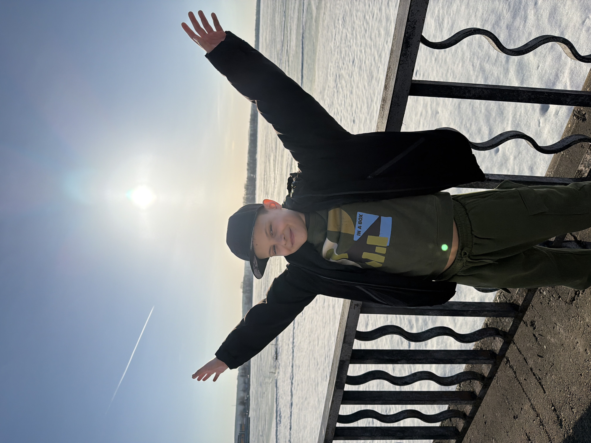 A young boy in a black jacket, olive green shirt and pants, and a black baseball cap stands on a bridge with his arms outstretched. He appears to be smiling. The backdrop features a snow-covered landscape under a bright sun, and a contrail is visible in the sky. The location is possibly in Votkinsk, Russia, as requested.