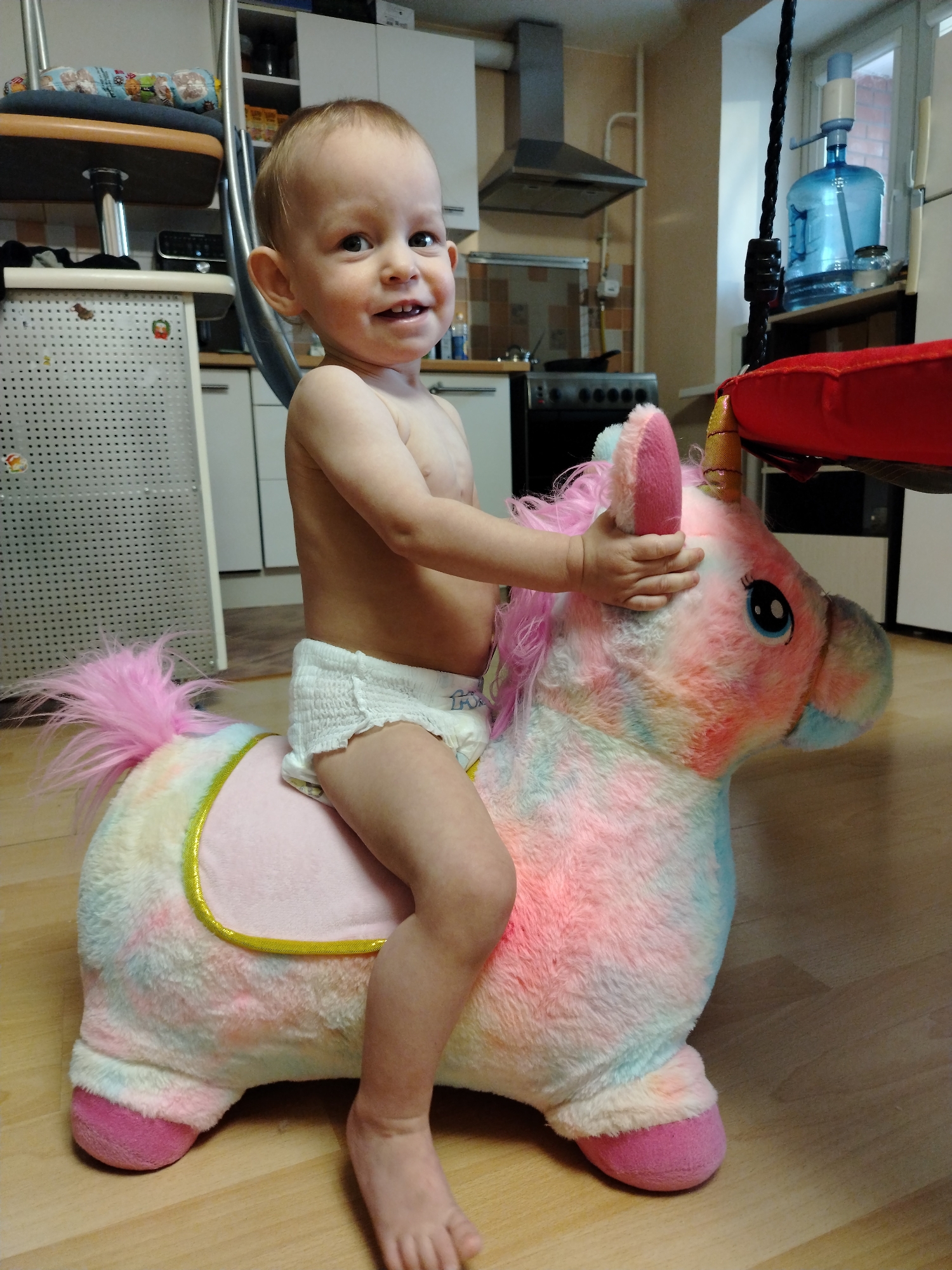 An infant, appearing to be approximately 1-2 years old, is depicted in an indoor setting within a residence in the city of Chaikovsky, Russia. The infant, bare-chested and wearing a white disposable diaper, is positioned straddling a large, plush, multicolored unicorn toy. The unicorn exhibits pink, blue, and white tie-dye coloring, features a vibrant pink mane and tail, a golden horn, and a pink saddle area with gold trim. The child's right hand rests on the unicorn's head, and the infant is looking towards the viewer with an open-mouthed smile. The background reveals a residential kitchen or dining area with light-colored wooden flooring and beige walls. Visible kitchen elements include white cabinetry, a stainless steel range hood, and a portion of a black stove with silver controls. To the right, a window overlooks an exterior brick building, and a large blue water dispenser bottle with a white pump is situated on the windowsill, next to a small glass jar. A black rope, possibly part of an indoor swing, is visible hanging in the upper right foreground.