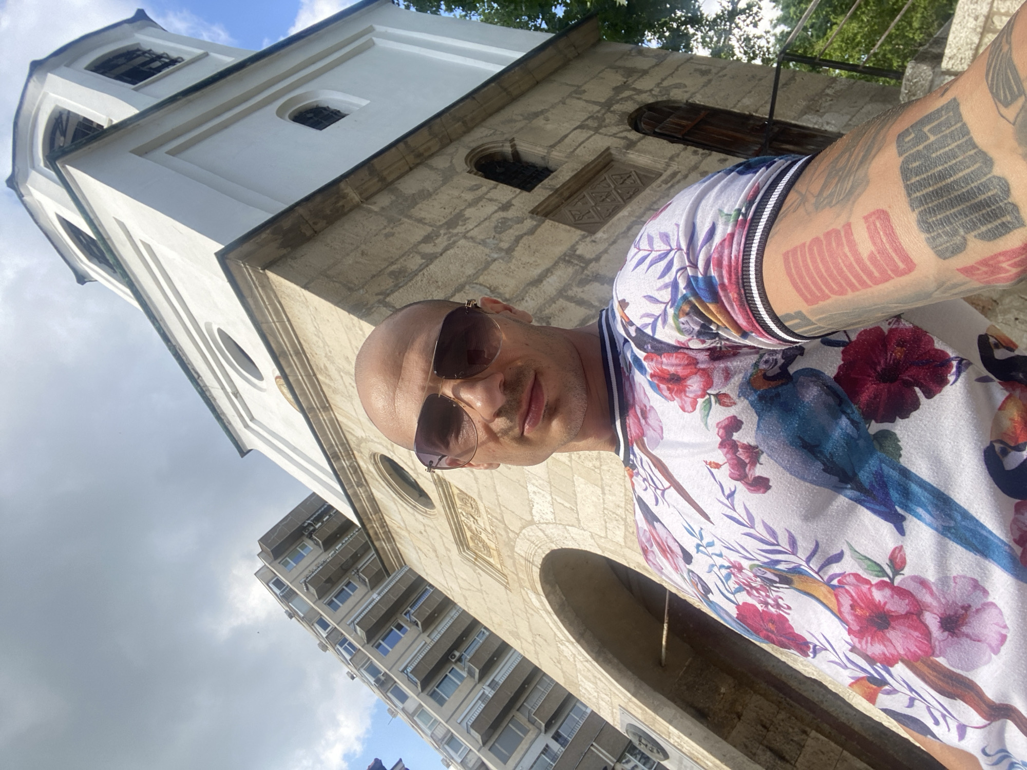 A low-angle selfie depicts a bald, light-skinned male with stubble, looking directly at the camera. He wears aviator-style sunglasses with brown reflective lenses and a white short-sleeved t-shirt patterned with colorful tropical birds (parrots, toucans) and pink-red flowers. His right forearm is visibly tattooed with block letter designs, including a red rectangular motif with horizontal lines and a black block design below it.
The scene is set in Pleven, Bulgaria. Behind the man, prominently featured, is the historic St. Nicholas Church (Църква Свети Николай). Its lower section is constructed from aged, light-colored, rough-hewn stone blocks, featuring a large archway and a circular window. Above this stone base rises a white, plastered bell tower with arched, dark-grille windows. To the left and behind the church, a modern, multi-story apartment building with numerous balconies and visible air conditioning units stands under a partly cloudy sky with patches of blue. Green foliage is discernible on the far right edge of the frame.