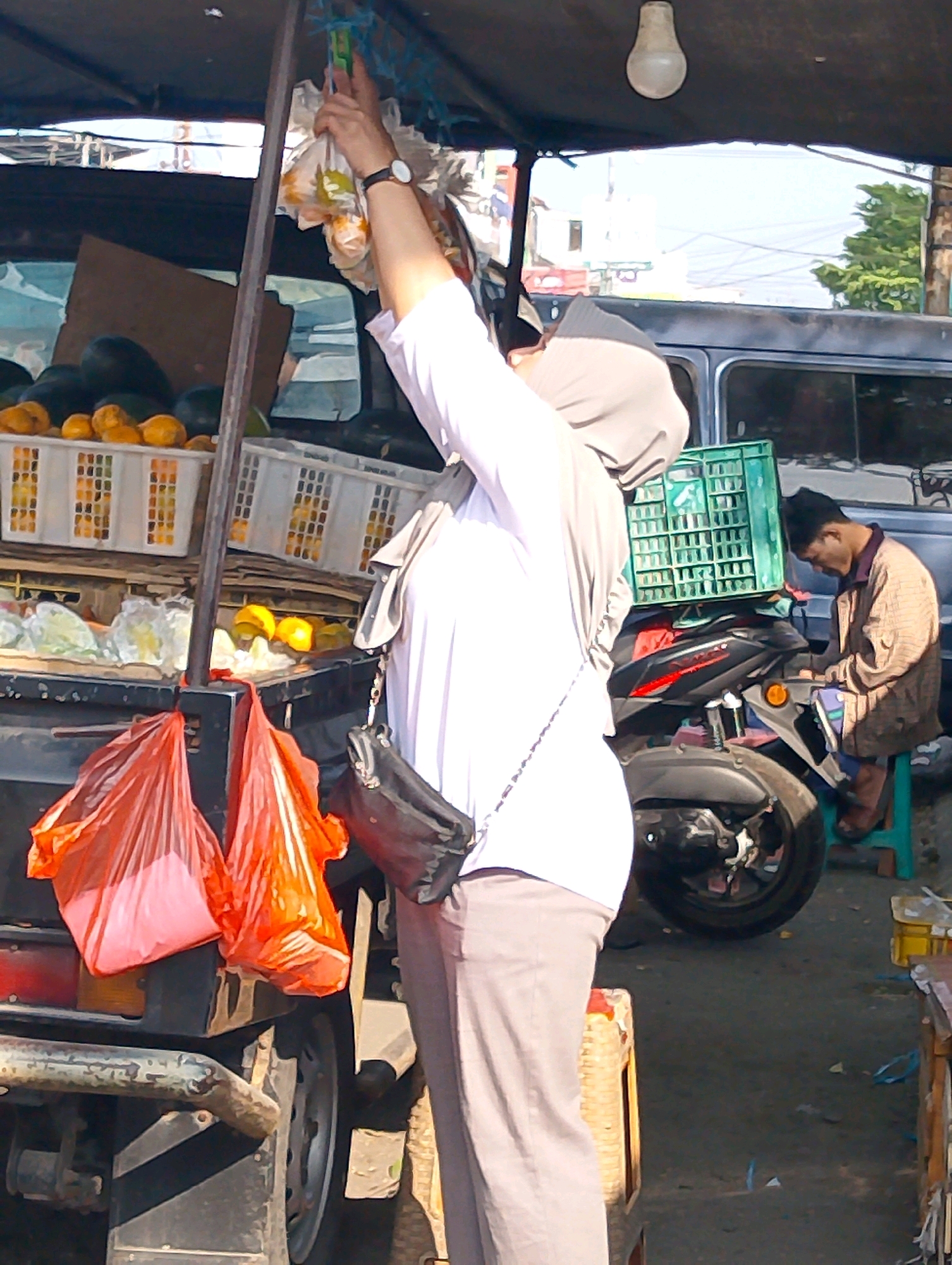 This image depicts a street scene in Lembang, Indonesia, likely near a market or roadside stall. In the foreground, a person wearing a light-colored headscarf and a white shirt with gray pants is seen from the side, reaching up to adjust items hanging from a makeshift structure, possibly a canopy or awning. The person is also wearing a dark shoulder bag.
Behind the person, there are several items for sale. White plastic crates filled with what appear to be fruits or vegetables are stacked. Two bright orange plastic bags hang from a part of the structure, obscuring the view of what they contain. A motorcycle is parked to the right, with a green plastic crate placed on top of it.
Further in the background, a man wearing a patterned shirt and dark pants is seated on a teal stool, looking down and appearing to be occupied with something. A van or large vehicle is visible behind the man and the motorcycle. The environment suggests an outdoor commercial setting, with a paved ground and the presence of trees and buildings in the distance, indicative of a town or city area. A light bulb hangs from the overhead structure. The lighting suggests it is daytime, with bright sunlight casting shadows.