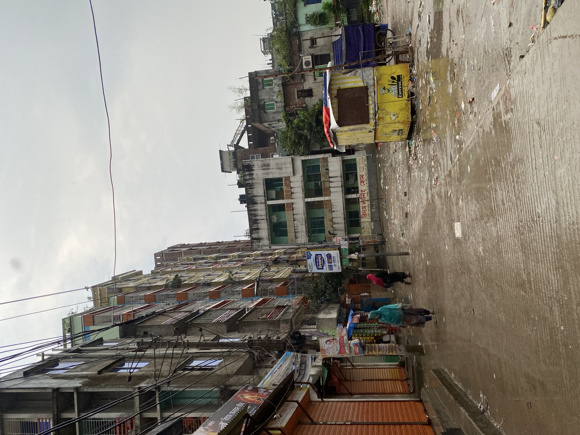 The image depicts a street scene in Dhaka, Bangladesh. The street is wet and littered with debris. Buildings line the street, with a mix of residential and commercial structures visible. Power lines are strung haphazardly across the view. Two people are walking down the street, and several stalls are set up along the roadside with closed metal shutters. A yellow food cart is parked further up the street. The sky is overcast. The overall impression is of a busy, somewhat chaotic urban environment.