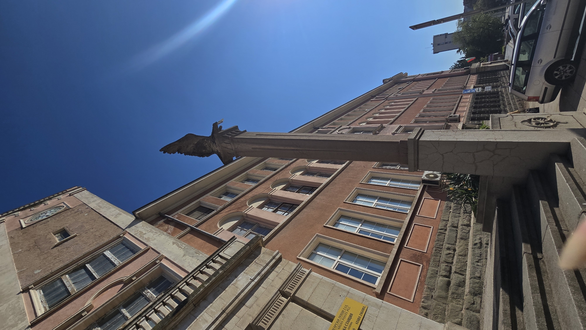 The image captures a view looking up at the architecture in Stara Zagora, Bulgaria. The scene features several buildings with reddish-brown facades and cream-colored detailing. One building has a clock on its facade. The sky is clear and bright blue. A large sculpture, possibly an eagle, projects from the top of one building. There is a stairway with a concrete railing in the foreground. A car is parked alongside the street. There is a yellow sign attached to a wall that contains Cyrillic text.