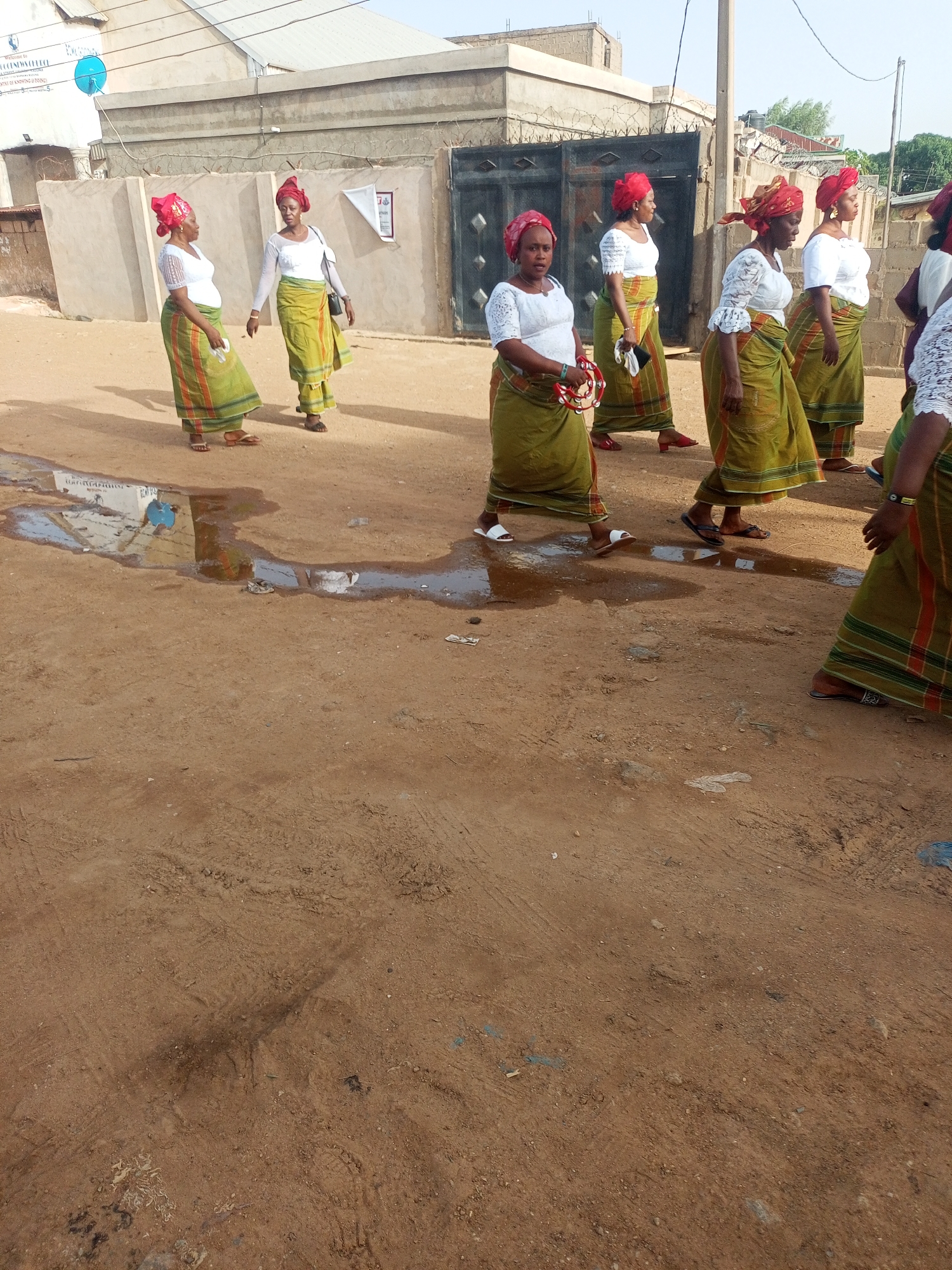 The image shows a group of approximately eight women walking in a line on an unpaved, reddish-brown dirt road in Sabon Tasha, Nigeria. They are uniformly dressed in cultural attire, consisting of bright red headwraps, white long-sleeved tops (many featuring lace or embroidered details), and long, olive-green wrap skirts with prominent red, orange, and darker green striped patterns near the bottom. Most of the women are wearing sandals or flip-flops.
The women appear to be moving in a procession or as part of a communal event. One woman in the foreground holds a small, round tambourine-like instrument, while another carries a black shoulder bag. Their expressions are generally calm and focused as they walk.
The setting includes several simple, single-story buildings made of concrete blocks in the background, some with corrugated metal roofs. A wall topped with barbed wire runs alongside the road, and a dark metal gate is visible behind some of the women. A blue satellite dish is mounted on one of the buildings. There are several puddles and wet patches on the dirt road, reflecting the sky and nearby structures.
The scene is illuminated by natural daylight, suggesting it is a sunny day, possibly late morning or early afternoon, with clear skies. Shadows are visible on the ground. On a white banner attached to one of the buildings in the background, text including "WELCOME TO COVENANT COLLEGE" is partially legible.
