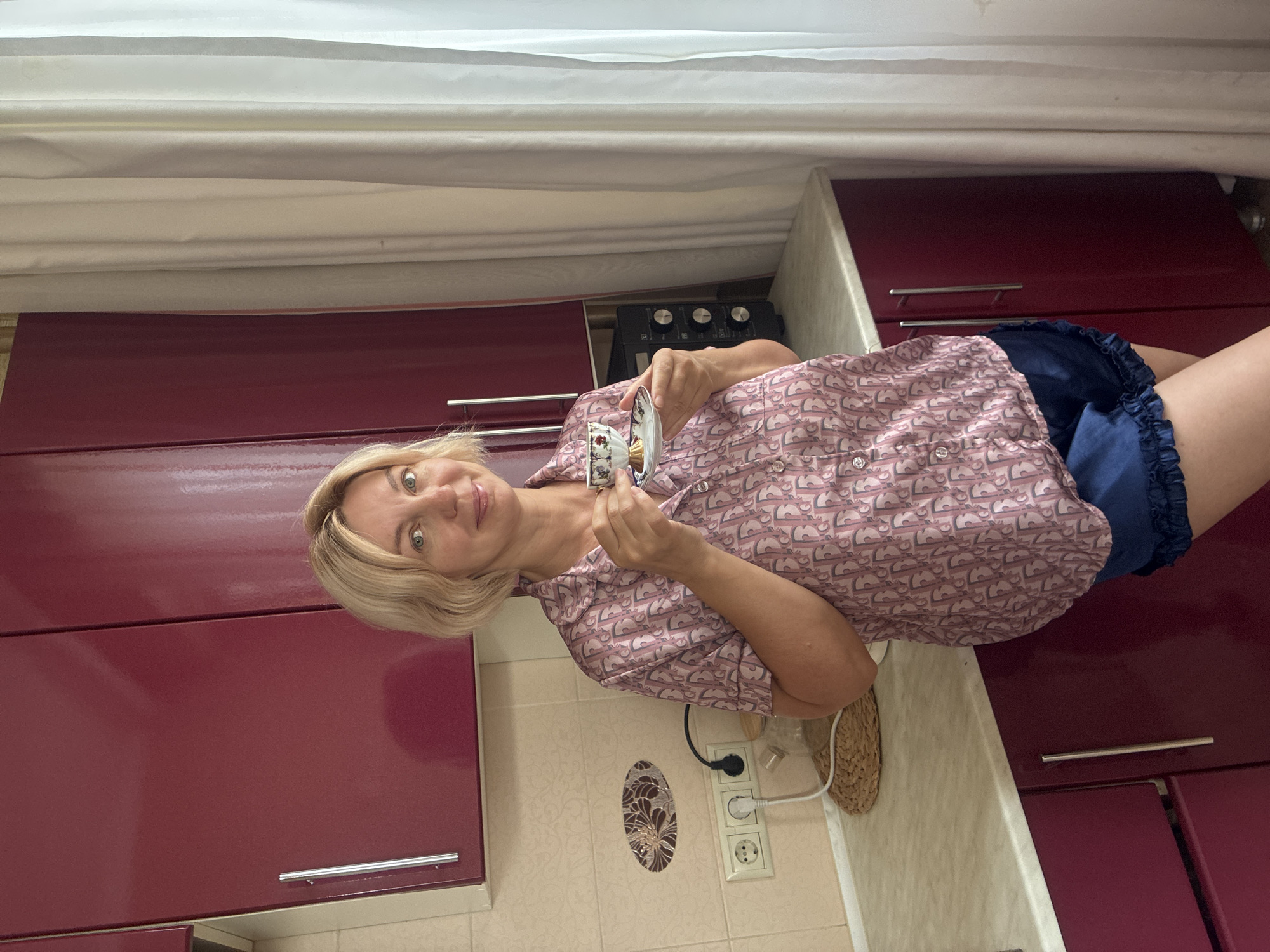 A blonde woman with blue eyes stands in a kitchen holding a teacup and saucer. She is wearing a pink patterned pajama top and dark blue shorts. The kitchen has dark red cabinets and light-colored countertops. There is a microwave on the counter behind her. To the right, white curtains hang by a window. The scene is indoors and appears to be a domestic setting.