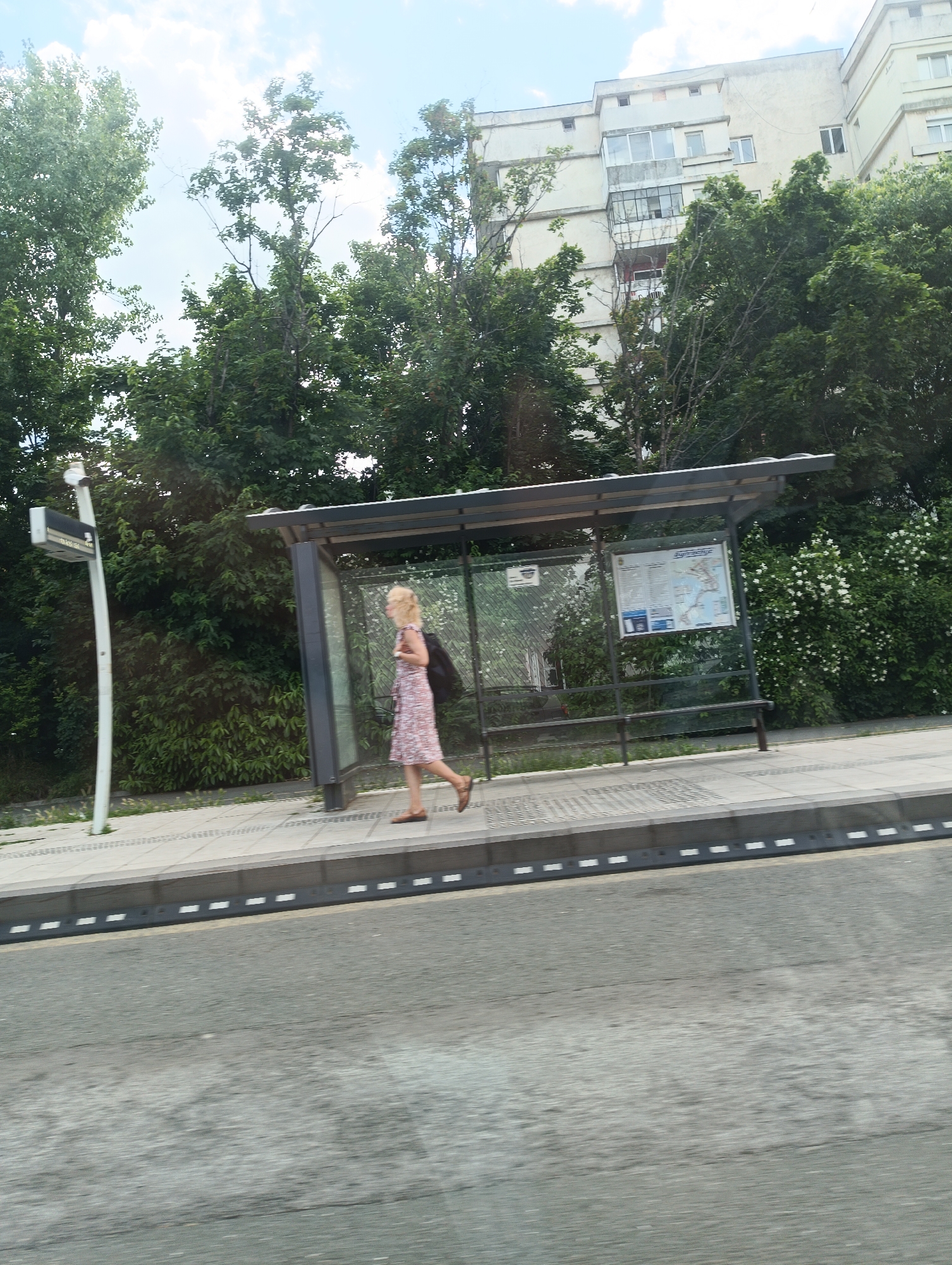 A woman with blonde hair wearing a floral dress and carrying a black bag walks past a bus stop in Burgas, Bulgaria. The bus stop has a roof, metal supports, and a bench. A bus schedule is displayed on the back wall. A street with a black and white striped curb runs in the foreground. Beyond the bus stop, there is a variety of trees and shrubbery. In the background, a multi-story residential building is visible through the trees. A street light is positioned on the sidewalk near the bus stop.
