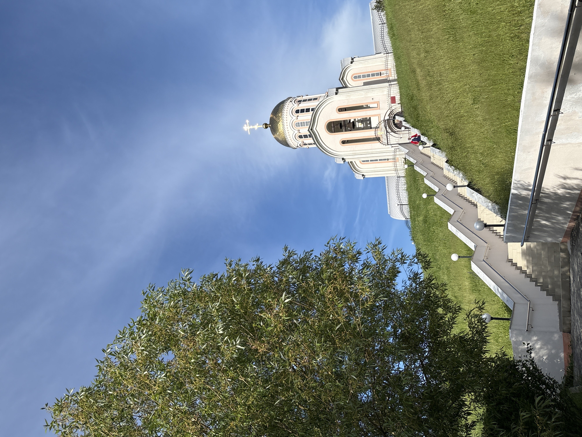 This image, when correctly oriented, captures a grand Orthodox church under a bright blue sky. The prominent central feature is the church building, characterized by its light-colored facade, numerous arched windows, and a magnificent golden dome crowned with a luminous white cross.
The church is situated at the top of a verdant, grassy hill. A wide, multi-tiered staircase with white marble steps and railings ascends the hillside towards the church entrance. Several spherical light fixtures are placed along the stairs, illuminating the path. In the foreground, a large, leafy green tree partially frames the lower left side of the scene, with its branches extending towards the center.
The sky is a clear, vibrant blue with only sparse, wispy clouds, indicating sunny and pleasant weather conditions. Shadows cast on the stairs and the ground suggest it is daytime, likely morning or afternoon. A few small, indistinct figures, possibly people, can be seen near the base of the church and at the top of the stairs; one figure appears to be wearing red.
The overall scene conveys a sense of grandeur and tranquility, characteristic of religious sites. The architectural style, particularly the golden dome and cross, strongly points to an Orthodox church, consistent with the provided location context of Varvarovka, Russia. There is no visible text in the image.