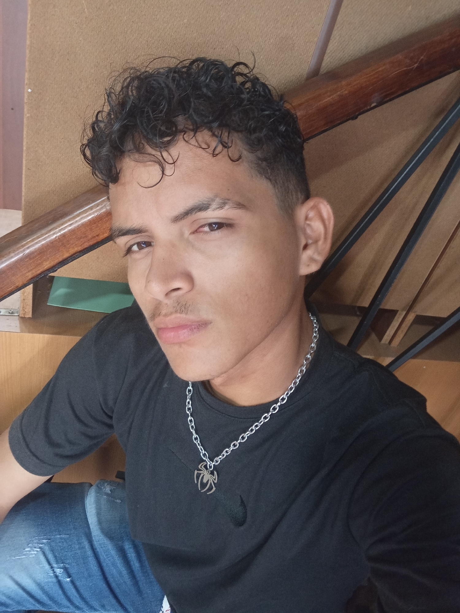 A young man with curly black hair, a mustache, and wearing a black t-shirt, jeans, and a silver chain with a spider pendant poses indoors. The background shows a wooden staircase and a light brown wall. The location is unidentified and cannot be determined to be Juan Griego, Venezuela from the image alone.