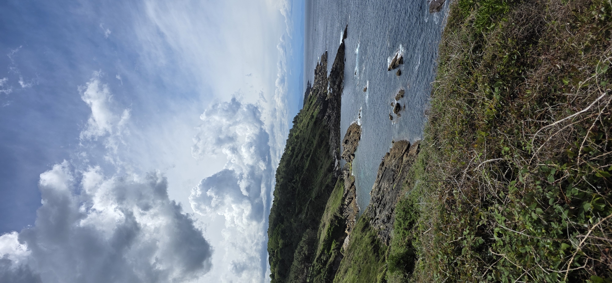 The image depicts a coastal scene in Hondarribia, Spain. From a high vantage point, the view extends over a rocky coastline and the ocean under a partly cloudy sky.
The terrain consists of a steep, green hillside with dense vegetation that transitions to rugged, dark rocks where the land meets the sea. Several rocks and small islands protrude from the water, with waves visible around them. The ocean water appears dark and textured.
The sky features a mix of blue sky and billowing white and gray clouds. The composition suggests a natural, outdoor environment with no visible human structures. There are no people visible in the image.