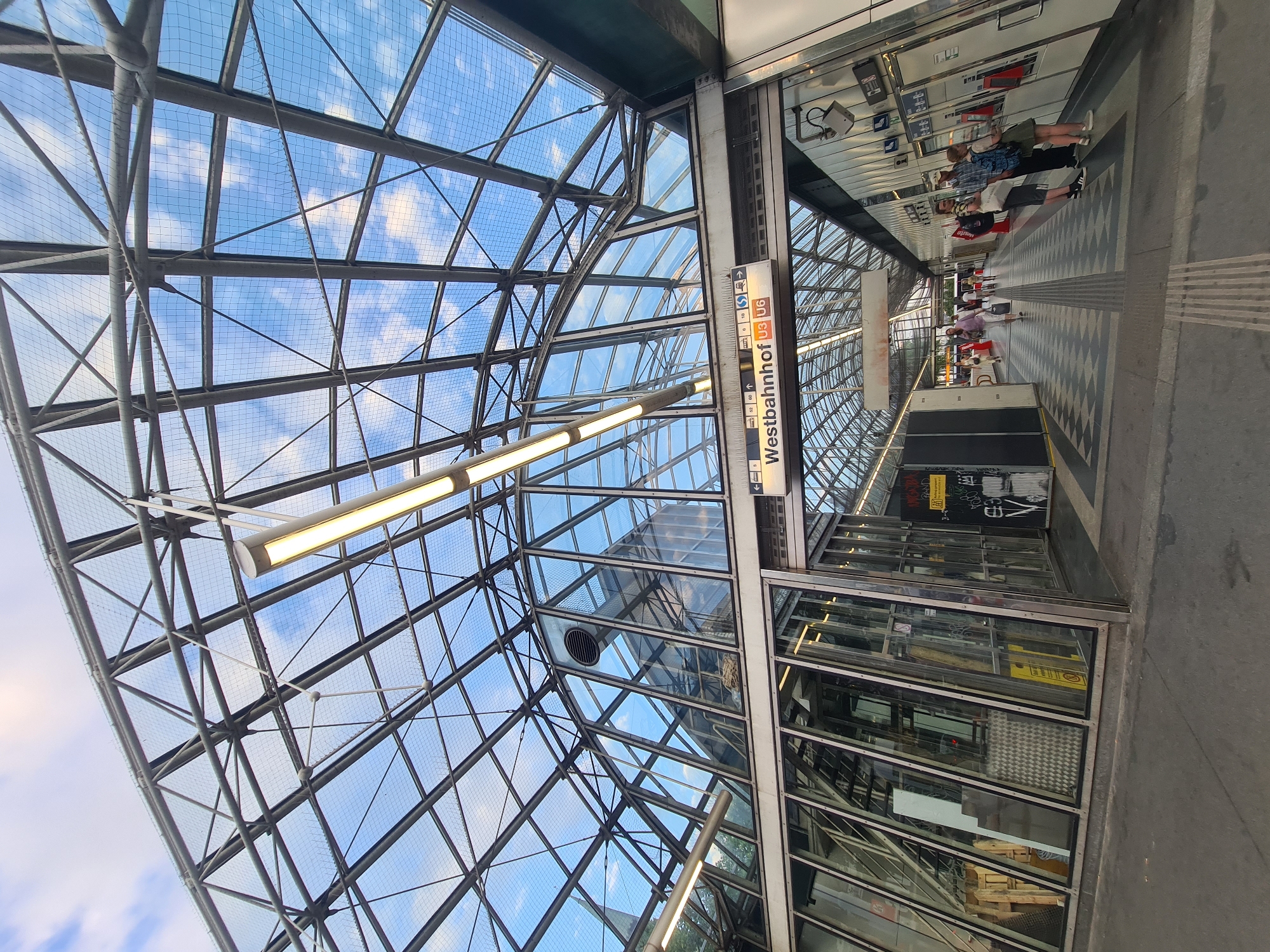 The image shows the interior of a train station with a glass and metal roof. The roof is supported by a complex metal structure and covered with netting. Sunlight streams through the glass, illuminating the station's interior, which features a tiled floor with a patterned design. A sign above an entrance reads "Westbahnhof U3 U6". Several people are visible, walking along the platform and standing near ticket machines. Some are carrying bags or wearing backpacks. To the left, there is an area with glass walls revealing stairs and what appears to be storage. The overall scene depicts a busy public transportation hub. The location is Vienna, Austria, specifically Westbahnhof station.