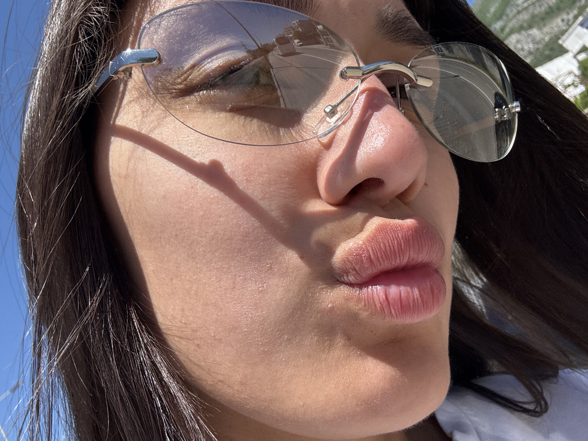 Close-up portrait of a female subject with dark hair, fair skin, and pursed lips. The subject is wearing rimless glasses with silver frames. The reflection in the glasses indicates an outdoor scene with a road and buildings. The location is possibly Zankovići, Montenegro, implied by the reflection. The lighting suggests a sunny day. The subject is facing away from the camera at a slight angle. The composition focuses on the subject's face, highlighting the lips, nose, and glasses.
