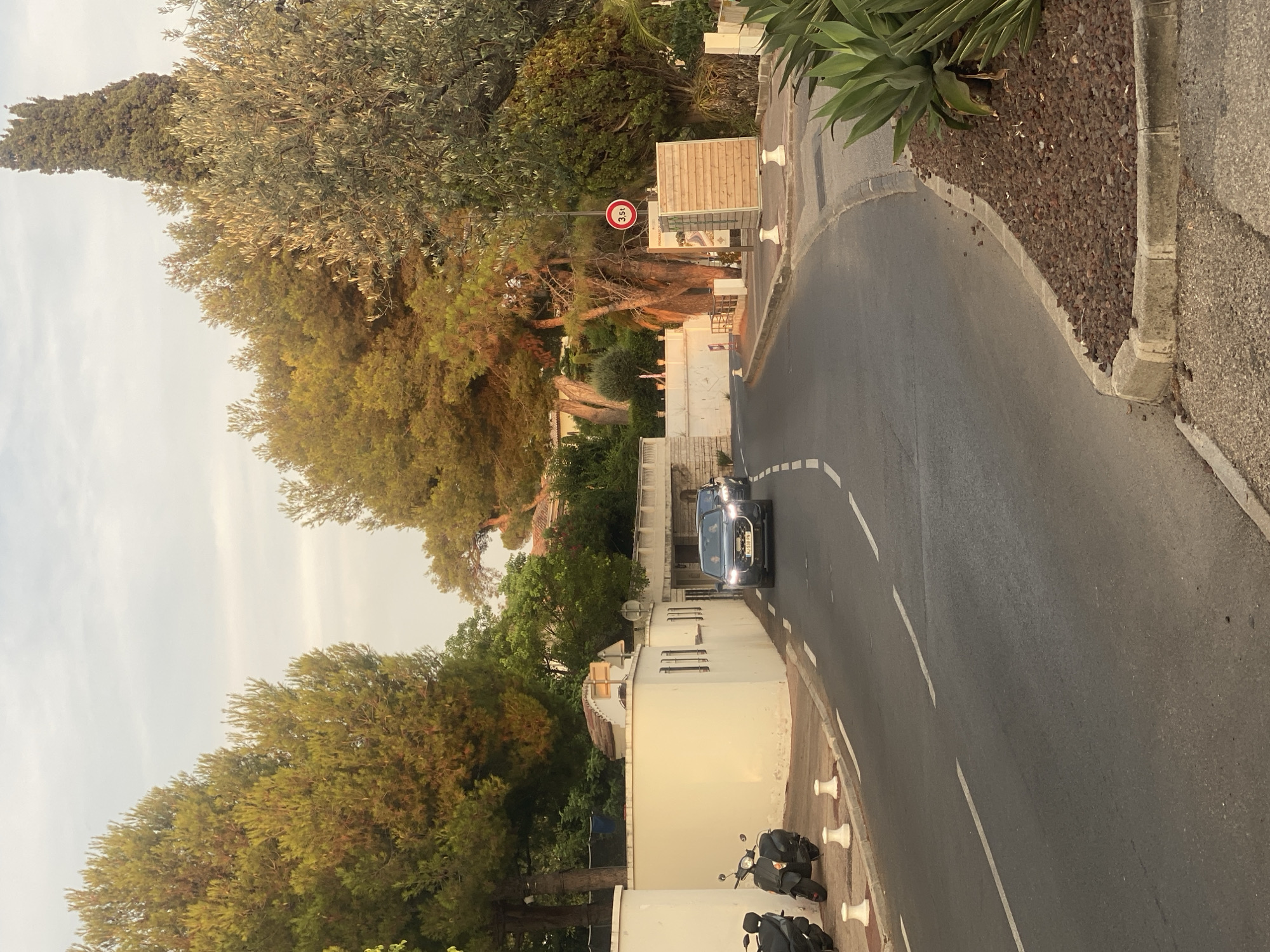 This image depicts a sunlit street scene in Antibes, France. A gray sedan with license plate "HJ 539 PN" is driving down a paved road. To the left of the road, there is a white wall with two parked dark-colored scooters. A white bollard is visible in front of the wall. Ahead on the right side of the road, a circular red and white sign indicates a weight limit of 3.5t. A wooden structure is positioned behind the sign. Lush green trees and foliage frame the scene, with a tall, slender cypress tree standing prominently in the background. The sky is partly cloudy.