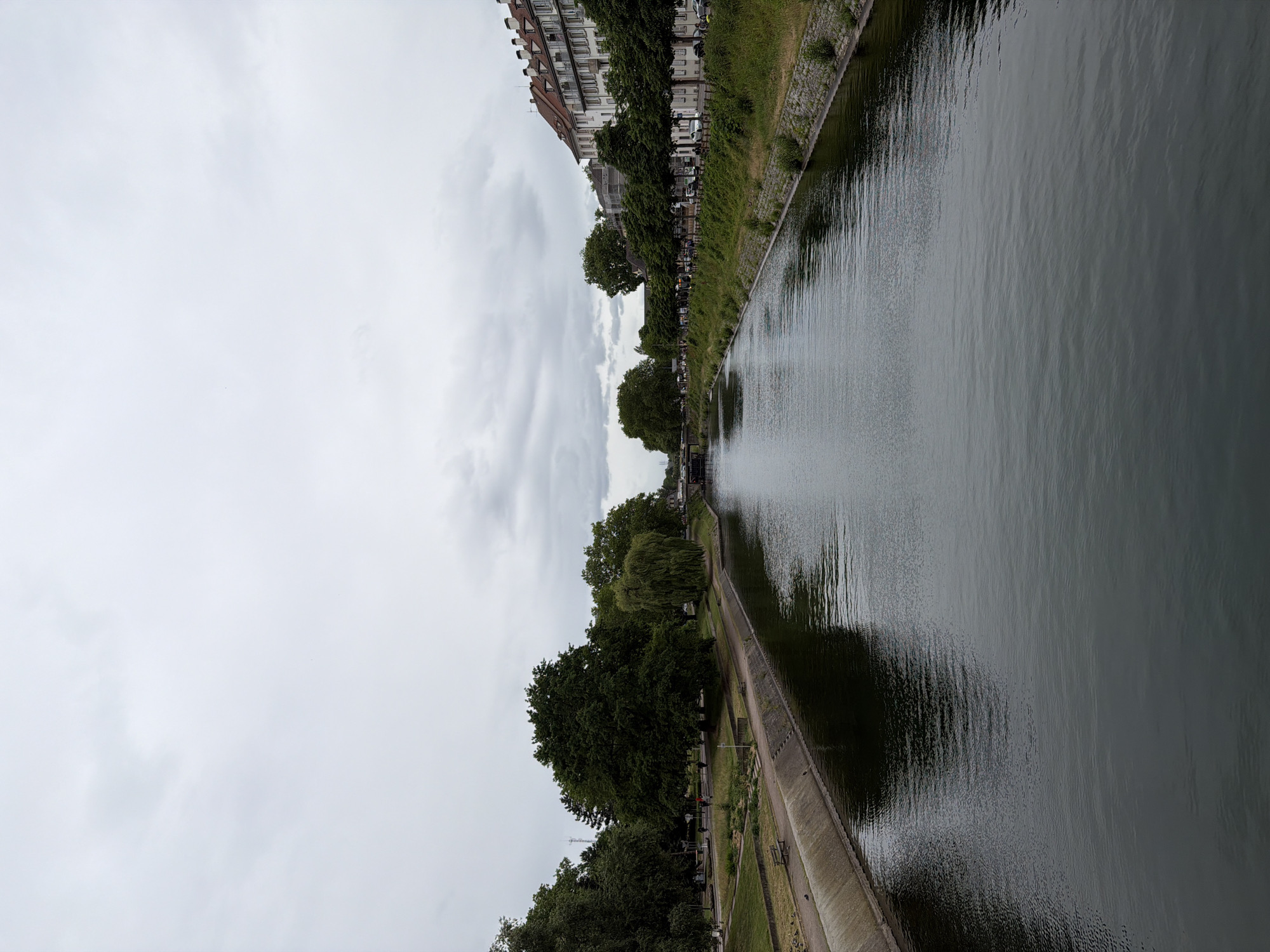 This landscape image captures a wide view of a river or canal in an urban setting, observed on an overcast day. The sky, dominating the upper left portion of the image, is filled with light grey clouds, indicating an absence of direct sunlight.
The river or canal occupies the right and lower sections of the frame. Its surface is calm but textured with subtle ripples, reflecting the cloudy sky and the trees lining its banks. Two distinct banks frame the waterway.
Along the left bank (when the image is correctly oriented), a row of multi-story buildings with classic European architecture is visible, featuring various window styles and some tiled roofs. A section of scaffolding is discernible on one of the buildings. A dense line of mature green trees partially obscures a road or pathway that runs alongside the water. Several vehicles, including cars and vans, are parked or moving on this thoroughfare, peeking through the foliage.
The right bank features a well-maintained concrete or stone-lined embankment, directly bordering a paved pedestrian path. Beyond the path are grassy areas and landscaped sections with small garden beds. A variety of trees, including a prominent weeping willow with trailing branches, are spaced along this bank, casting reflections onto the water. Several people are visible strolling along the path; one individual in a red top stands out. In the distance on this side, benches, a lamppost, and a tall mast-like structure can be seen.
The scene depicts a typical day in a city, with people engaging in leisurely walks by the water. The weather appears cool and subdued due to the heavy cloud cover. Given the location context, this is a view of Strasbourg, France. No discernible text is visible in the image.