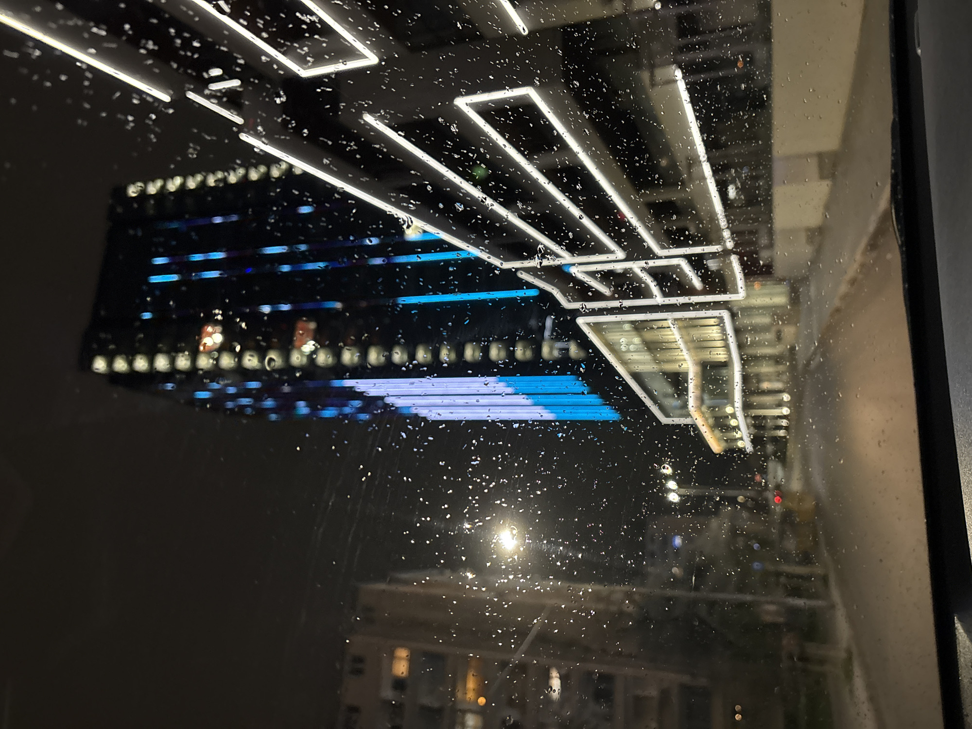 The image is a nighttime view of Sofia, Bulgaria, seen through a rain-streaked window. The reflection of a modern building with illuminated white rectangular accents and blue horizontal bands is prominent in the upper portion of the image. Below the reflection, a blurred view of another building with lit windows can be seen. The environment suggests it is nighttime and raining. There are no visible people, actions, or interactions directly depicted. The reflection of the building is likely the Sofia Tower, and the view through the window is of the surrounding city.