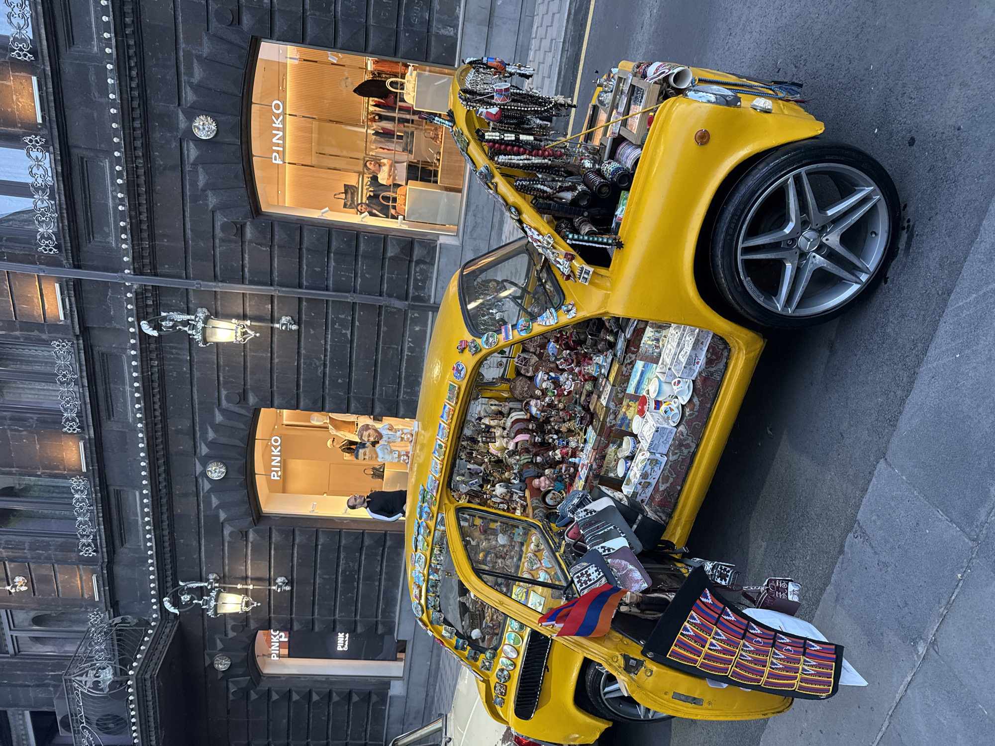 The image shows a yellow car parked on a street in Yerevan, Armenia. The car is heavily decorated inside and out with souvenirs, including mugs, dolls, badges, carpets, and an Armenian flag, suggesting it's a mobile souvenir shop. The car's hood is open, displaying more souvenirs, primarily jewelry. The background shows a building with two "Pinko" storefronts, indicating a retail area. The lighting suggests it's daytime, likely early evening. A person in a black jacket can be seen in one of the storefronts. The scene appears to be a tourist area with the car functioning as a vendor.