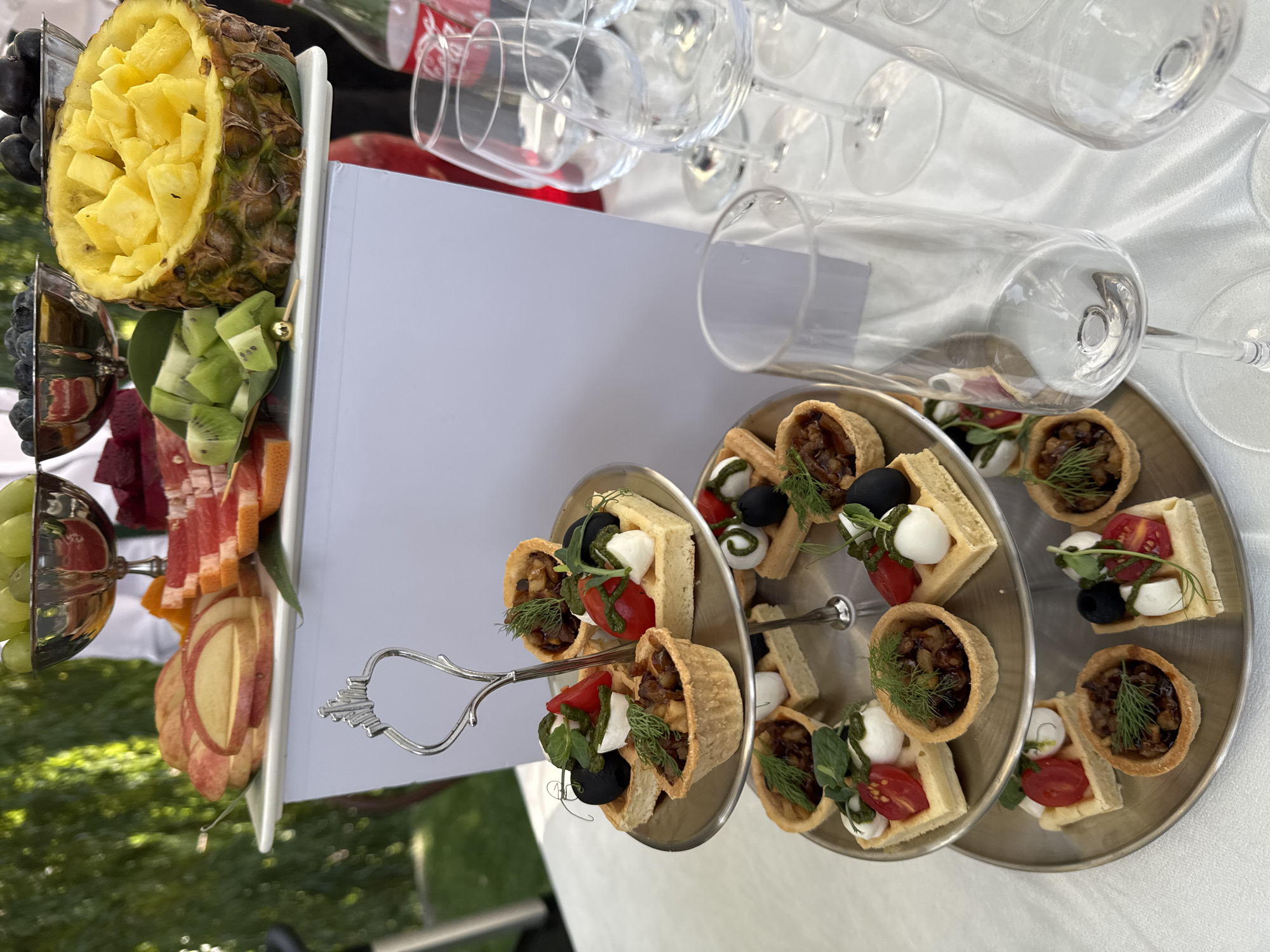 A three-tiered silver serving tray holds an assortment of appetizers, each adorned with small garnishes. The appetizers appear to be small tartlets and square waffle-like bites topped with ingredients such as cherry tomatoes, mozzarella balls, olives, and diced meat or vegetables. Above the serving tray, a white rectangular structure displays a variety of fruits including sliced apples, grapefruit, kiwi, grapes, and pineapple carved into a bowl shape. The scene is set outdoors, with lush green foliage visible in the background, suggesting a garden or park setting. Several empty wine glasses and bottles of Coca-Cola are arranged on the white tablecloth in the foreground. The overall ambiance suggests a catered event or a festive gathering. The image was captured in Koy Tash, Kyrgyzstan.