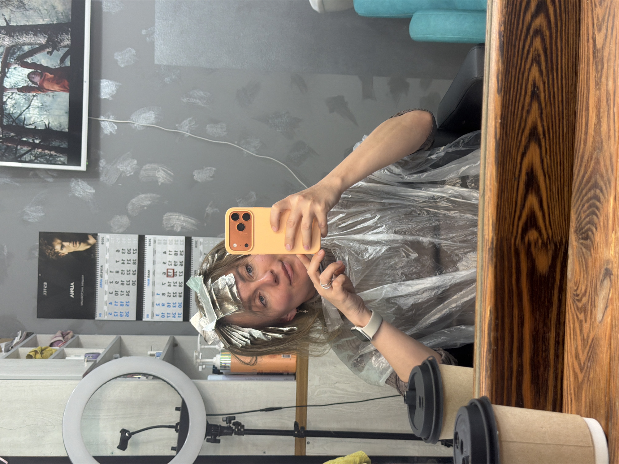 A woman with foils in her hair is taking a selfie in what appears to be a hair salon. She is wearing a plastic cape, holding a yellow smartphone up to capture her reflection in a large mirror. The reflection shows a ring light, salon products, and a calendar with dates visible (possibly including '3034', '3036', '2034', '2036'). The presence of the calendar, salon products, and hairstyling foils suggests that this is happening in a professional salon setting. A framed picture hangs on the gray wall, and a wooden frame runs along the side, partially blocking the view. Based on the calendar in the background, the image could potentially be related to or from Chaikovsky, Russia, given that 'Чайковский' appears written in Cyrillic next to the calendar's dates.
