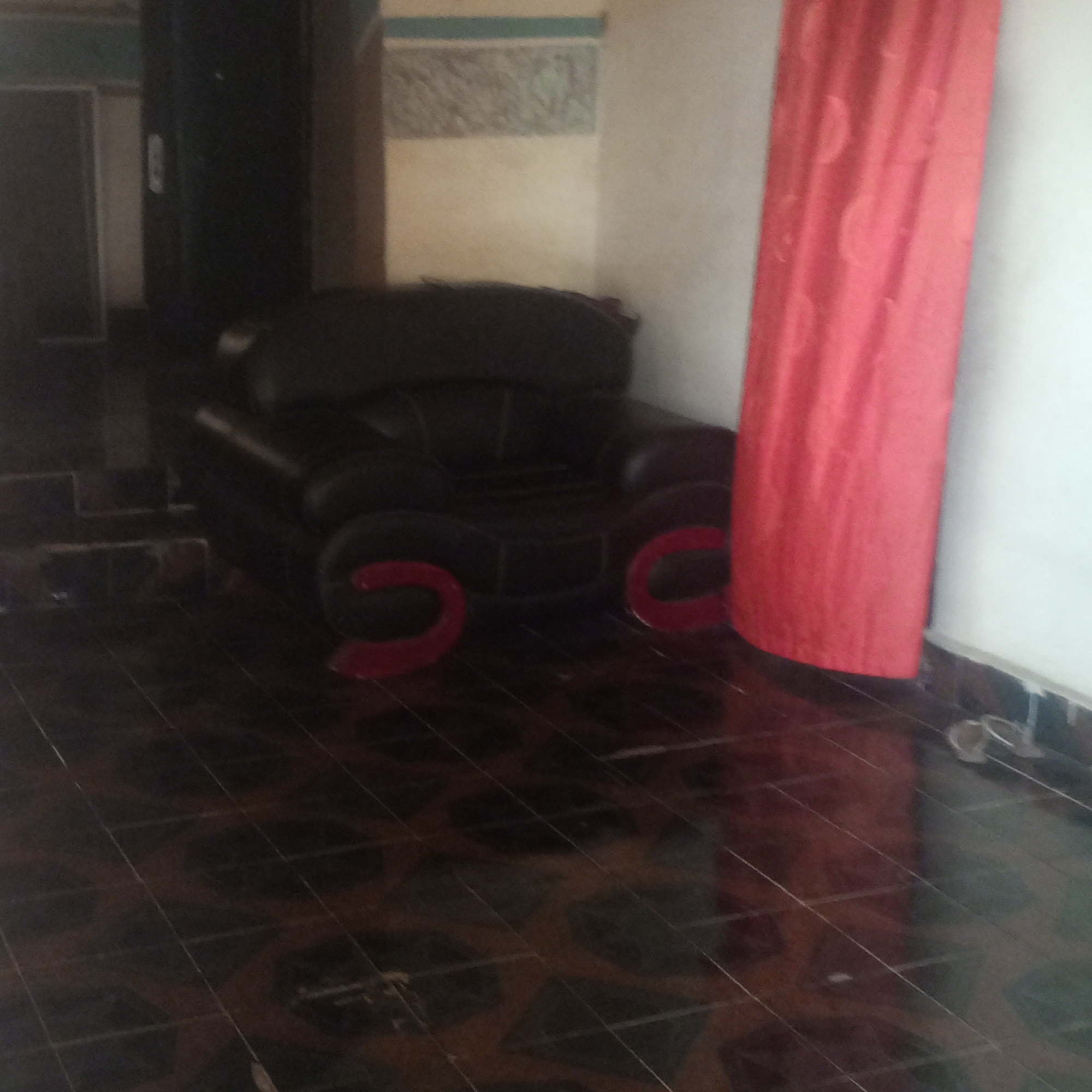 In an indoor scene at an unknown location, a black leather armchair sits on a dark, glossy tiled floor. The chair has a curved design with a wide seat and arms. Two red, curved objects are placed on the floor in front of the chair, one on each side. To the right of the chair, a bright red curtain hangs vertically, partially obscuring the wall. The wall behind the curtain is white, and further to the left, there is a darker wall or door. The background features patterned wallpaper. The lighting is dim, casting shadows on the floor. A pair of light-colored flip-flops is visible near the bottom right corner of the image.