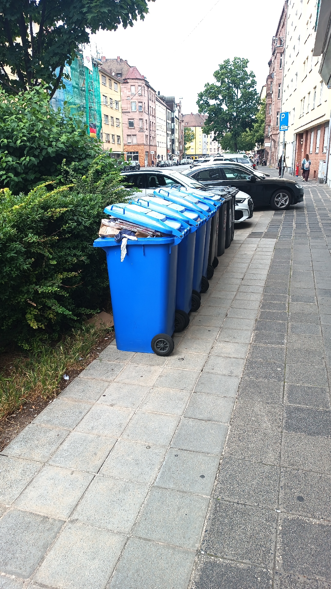 The image depicts a row of blue garbage bins on a sidewalk in Nuremberg, Germany. The bins are made of plastic and have wheels. The first bin is open and contains some paper and fabric. Behind the bins, several cars are parked along the street. In the background, there are buildings with windows and balconies. A green bush is visible on the left side of the image. The sidewalk is paved with square tiles. A parking sign is visible on the right side of the image. The scene appears to be a typical urban street environment.