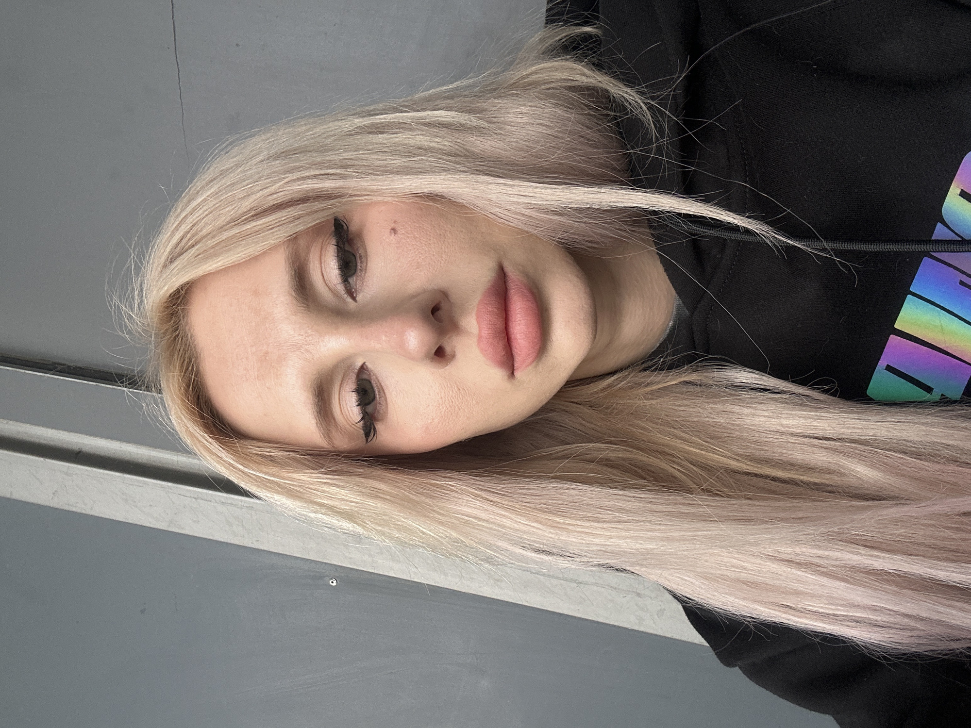 A selfie of a woman with long blonde hair and pink lips, wearing black eyeliner. She is wearing a black hoodie. The background is a gray wall.