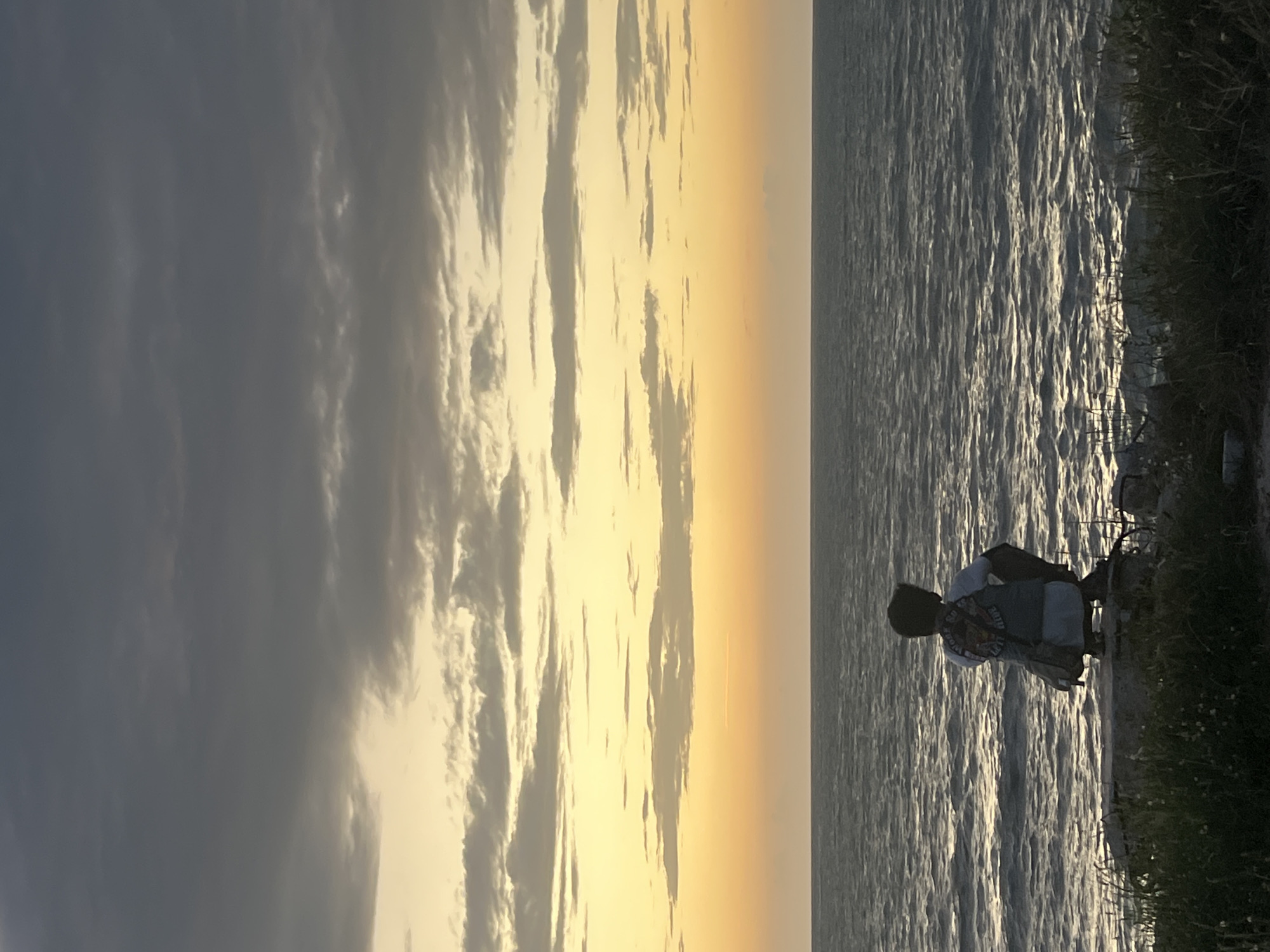 A lone figure is seen crouching on a rocky outcrop overlooking the ocean, facing away from the camera. The individual, wearing a backpack, is silhouetted against a dramatic sky with layers of clouds. The sky transitions from a deep grey at the top to soft yellow and orange hues near the horizon, suggesting either sunrise or sunset. The ocean stretches out to the horizon, its surface rippling with reflected light. The foreground is partially obscured by grass and vegetation. The overall impression is one of quiet contemplation in a natural setting. The location is described as Ko Pha-ngan District, Thailand.
