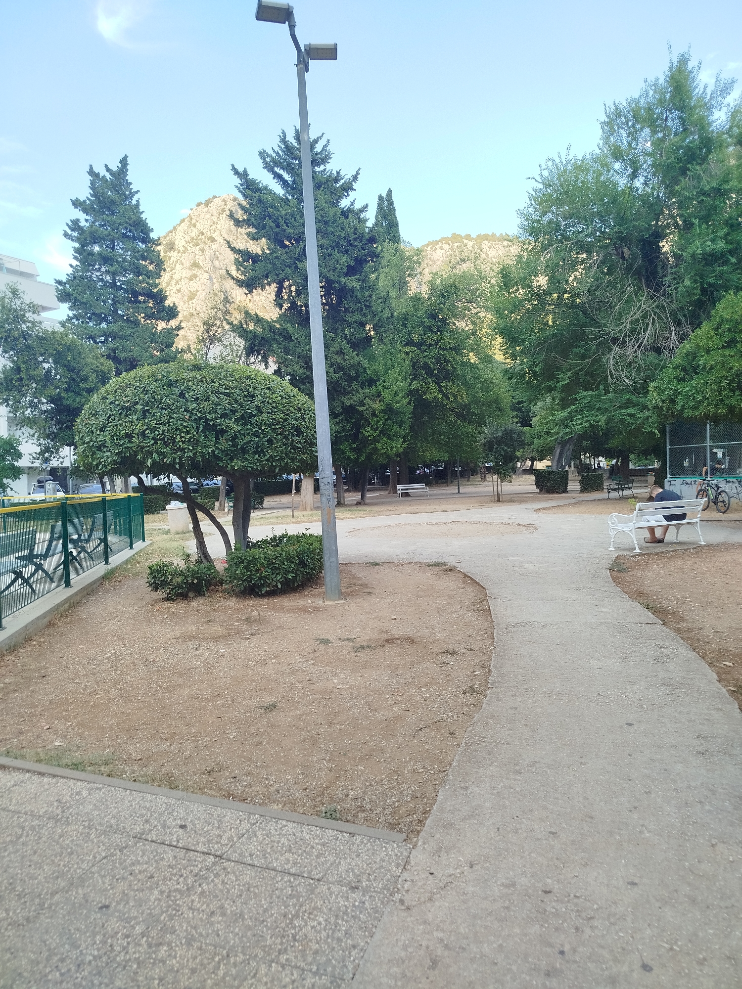 The image depicts a park scene in Omiš, Croatia, with a prominent lamppost in the foreground. A paved walkway curves through the park, bordered by gravel and low-lying bushes. In the background, several trees are visible, some with rounded canopies, and behind them, a rocky mountain slopes upward. A white bench is occupied by a person sitting with their back to the camera, and further in the distance, another white bench is visible with no occupants. A green fence runs along the left side of the image, with a building peeking out from behind the trees. The sky is clear and blue.