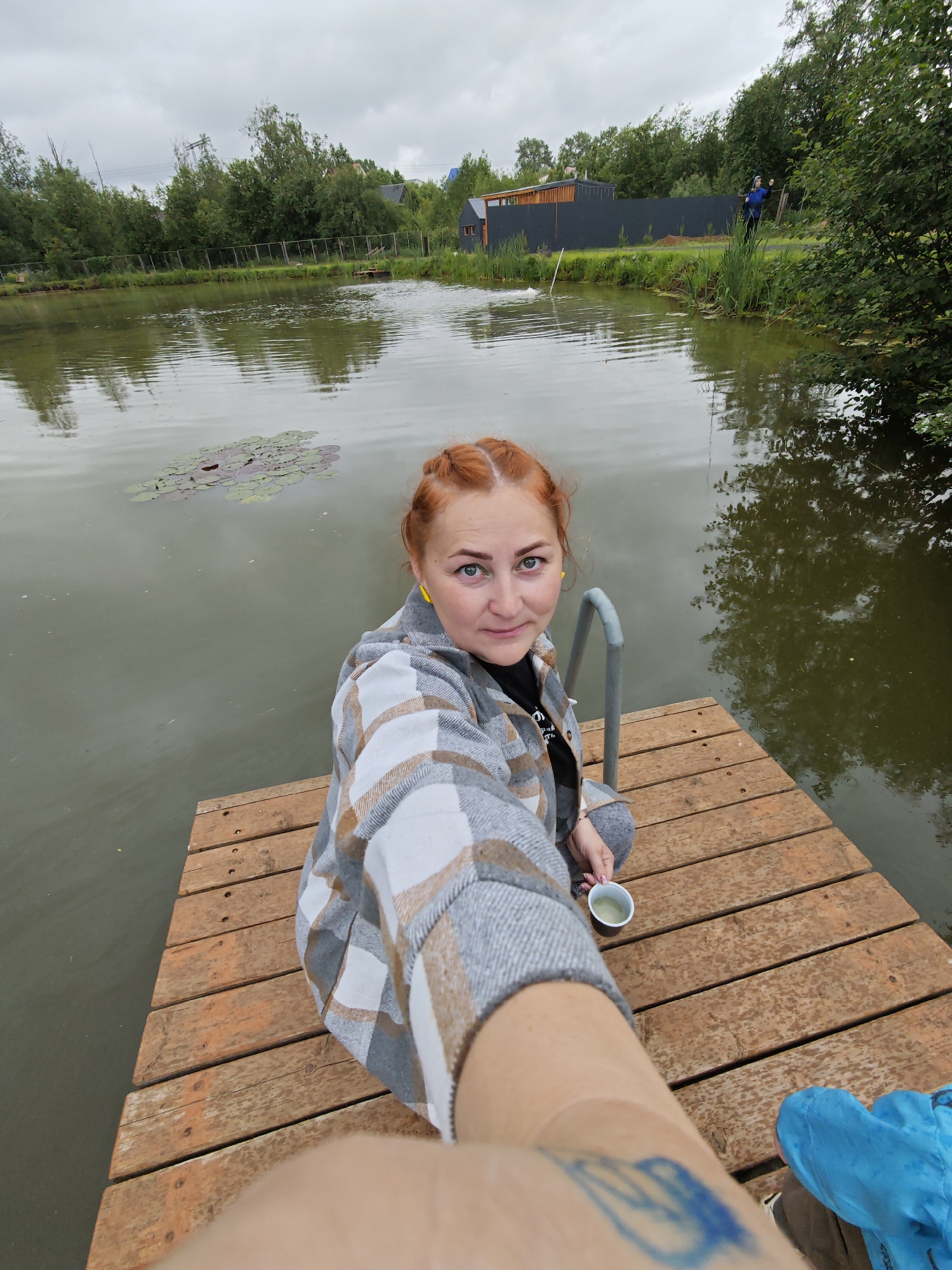 The image captures a selfie of a woman sitting on a wooden dock beside a pond in Malyi Bukor, Russia, under an overcast sky. The woman has reddish hair styled in two top buns and light-colored eyes, looking directly at the camera with a neutral expression. She wears a plaid jacket in shades of grey, white, and tan over a dark t-shirt and grey pants. A yellow earbud is visible in her left ear. She holds a small white cup containing a light green liquid in her left hand. Her right forearm is extended towards the camera, showing a partially visible blue design.
The dock is constructed from reddish-brown wooden planks and features a silver-grey metal handrail to the woman's right. The pond's water is a murky olive-green, reflecting the cloudy sky and surrounding foliage, with gentle ripples on its surface. Several lily pads, some with reddish hues, float on the water near the dock.
The far bank of the pond is lined with dense green trees. Behind the trees, a dark fence is visible, alongside a long, dark-colored building with lighter wooden accents. A smaller, dark structure is situated further to the right. A person wearing a blue top stands on the far bank, partially obscured by the trees, appearing to engage in an activity by the water. Utility poles and lines are discernible above the distant treeline.