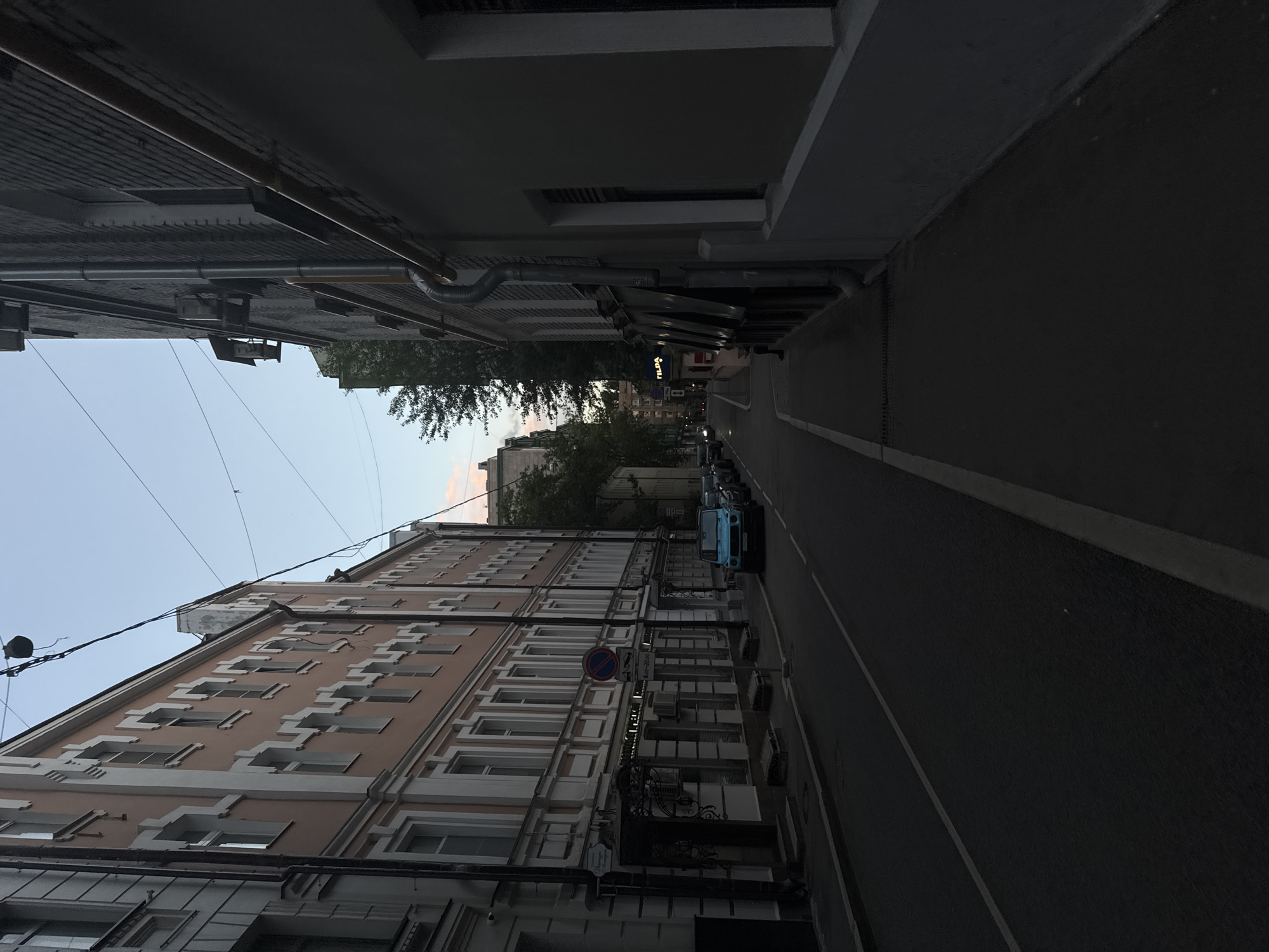 The image depicts a narrow street in Moscow, Russia, at dusk. On the left, a light pink building with white decorative trim lines the street, featuring multiple windows. A darker, taller building with visible pipes and a grey facade stands on the right. The street itself is paved, with a white line marking the lane. Several cars are parked along the right side of the street, and a bright blue car is visible further down on the left. Trees are present in the background, peeking over the buildings. Power lines crisscross the sky, which is a pale blue with hints of pink from the setting sun. The overall scene conveys a quiet urban atmosphere.