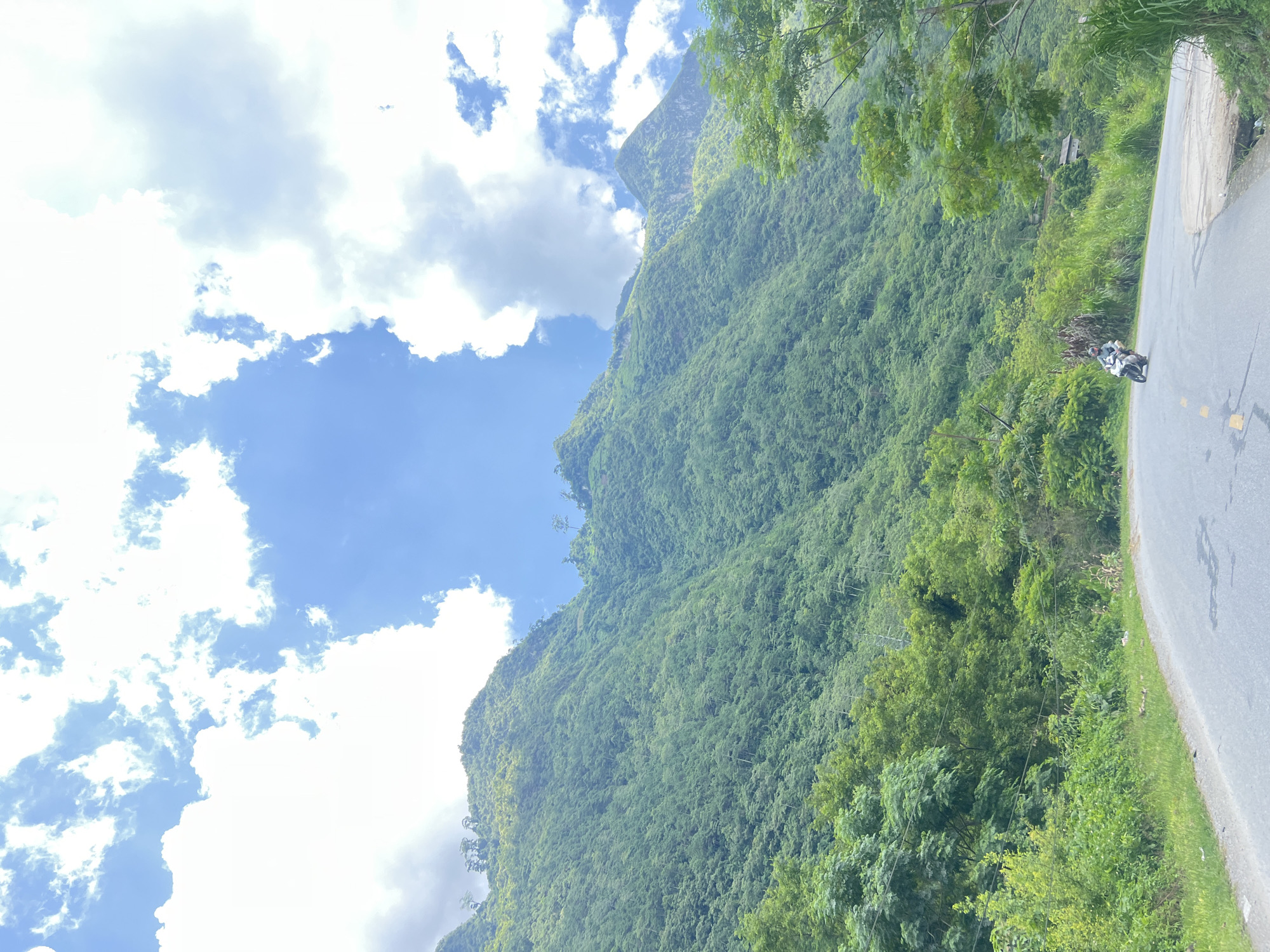 A paved road curves through a lush green landscape with steep, tree-covered mountains in the background. The sky is a vibrant blue, dotted with fluffy white clouds. A lone motorcyclist is visible on the road, riding towards the viewer. The scene appears to be in Minh Tân Commune, Vietnam, based on the provided information. The vegetation is dense and verdant, suggesting a tropical or subtropical climate. The road is empty except for the motorcyclist, and there are no other visible signs of human activity or structures. The overall impression is one of natural beauty and a serene, remote setting.