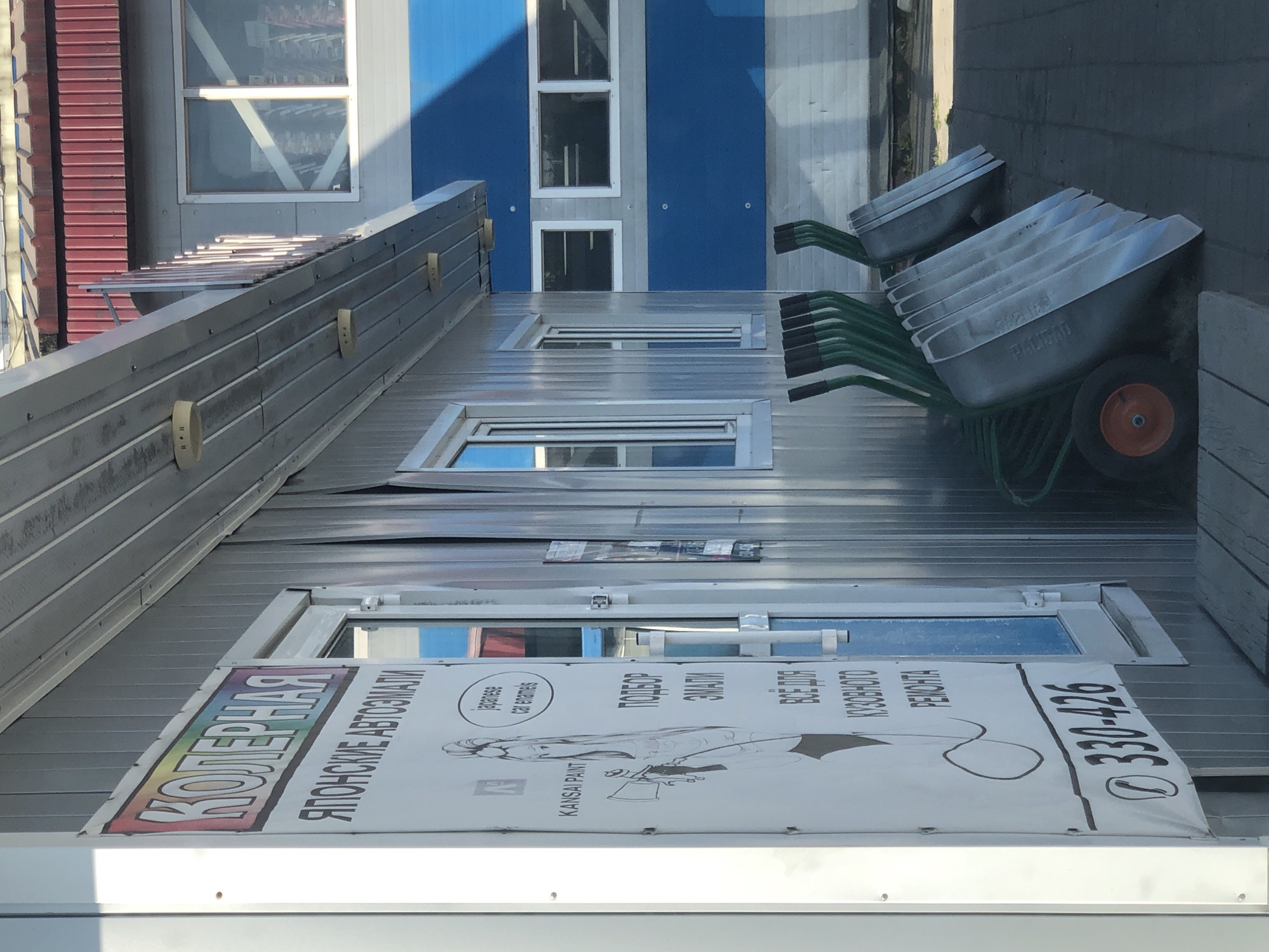The image captures the exterior of a small, likely commercial building, possibly a kiosk or workshop, in Petropavlovsk-Kamchatsky, Russia. The building features a corrugated metal facade, with a prominent sign on the left side. The sign, with a rainbow-colored header that reads "КОЛЕРНАЯ" (Color Shop), advertises "ЯПОНСКИЕ АВТОЭМАЛИ" (Japanese Auto Enamels). Below this, there's a graphic of a person holding a spray gun, along with text indicating "KANSai PAINT," "ПОДБОР ЭМАЛИ" (Enamel Selection), "ВСЕ ДЛЯ КУЗОВНОГО РЕМОНТА" (Everything for Body Repair), and a phone number, "330-426".
Adjacent to the building, a row of wheelbarrows is neatly arranged, with their metal basins facing outwards. Some of the wheelbarrows are stacked on top of each other. The ground in front of the building appears to be paved with interlocking bricks, suggesting an outdoor service or storage area. In the background, a larger building with blue and gray sections and windows is visible. The overall scene suggests a business operation related to automotive painting and repair, situated in an urban or semi-urban setting.