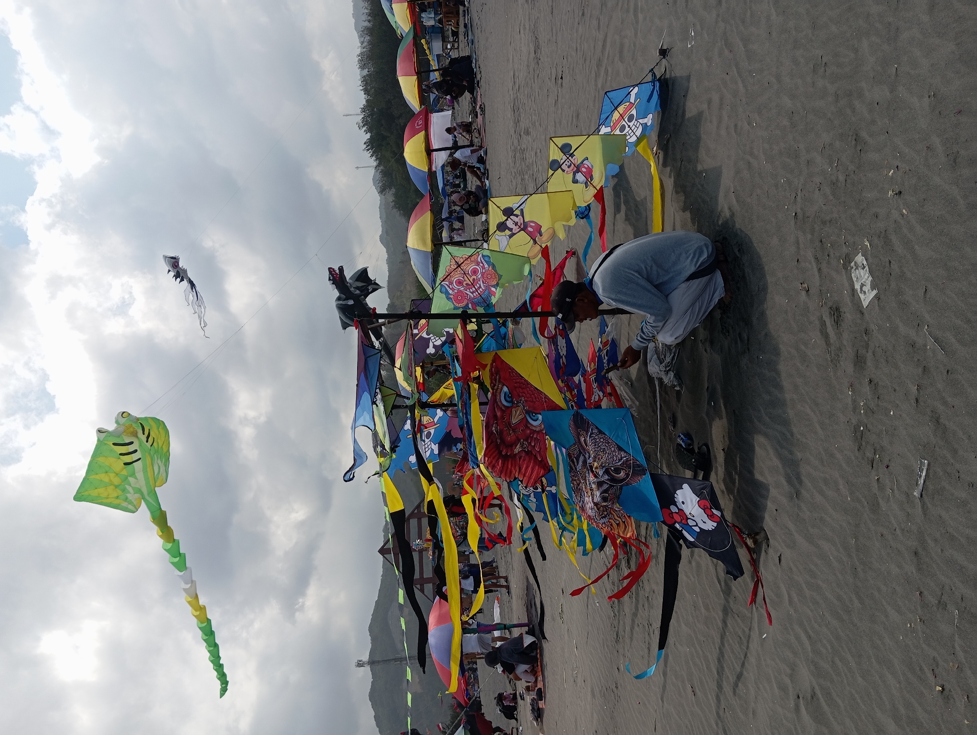 A man is kneeling on a sandy beach in Parangtritis, Indonesia, in front of a collection of colorful kites displayed on poles. The kites, featuring various designs including cartoon characters and traditional motifs, are suspended in the air and on the ground, with some trailing long ribbons. Several beach umbrellas are set up behind the kites, providing shade for people sitting on the sand. In the background, a gentle slope of hills covered in trees can be seen under a cloudy sky. The scene captures a lively atmosphere, possibly during a kite festival or a day at the beach where kite flying is a popular activity.