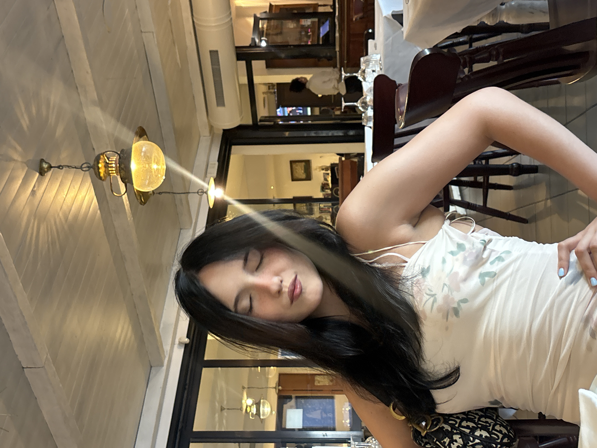 A young woman with long, dark hair and her eyes closed is seated at a table in a restaurant. She is wearing a white dress with a floral print and a black and white patterned purse is on her left shoulder. Her right hand is resting on her hip, and her nails are painted light blue. The restaurant has a white, slatted ceiling with pendant lights. Tables are covered with white tablecloths and set with glassware. In the background, through an open doorway, another person wearing a light-colored shirt is visible. The scene suggests a relaxed meal in a dining establishment. The image was taken in San Benedetto del Tronto, Italy.
