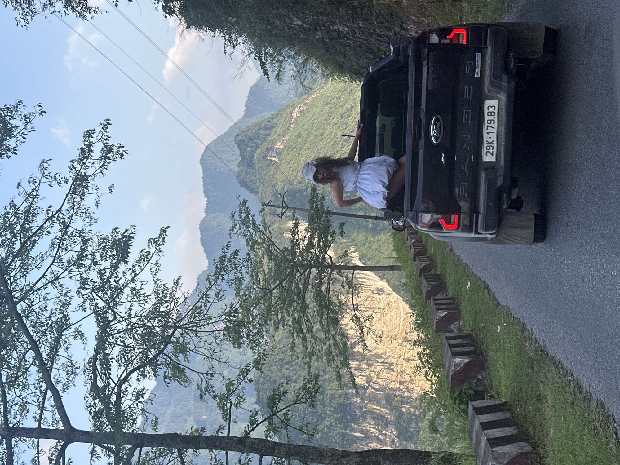 The image captures a scenic view in Đường Thượng Commune, Vietnam. A black Ford Ranger pickup truck is parked on the edge of a road with a guardrail. The truck's license plate reads "29K-179.83." A person, likely a woman, is positioned inside the truck bed, dressed in a white dress and a white head covering. Mountains covered in lush greenery form a backdrop. The sky is partly cloudy. Power lines are visible in the background. The road runs along a cliff overlooking a valley. Trees are present in the foreground.