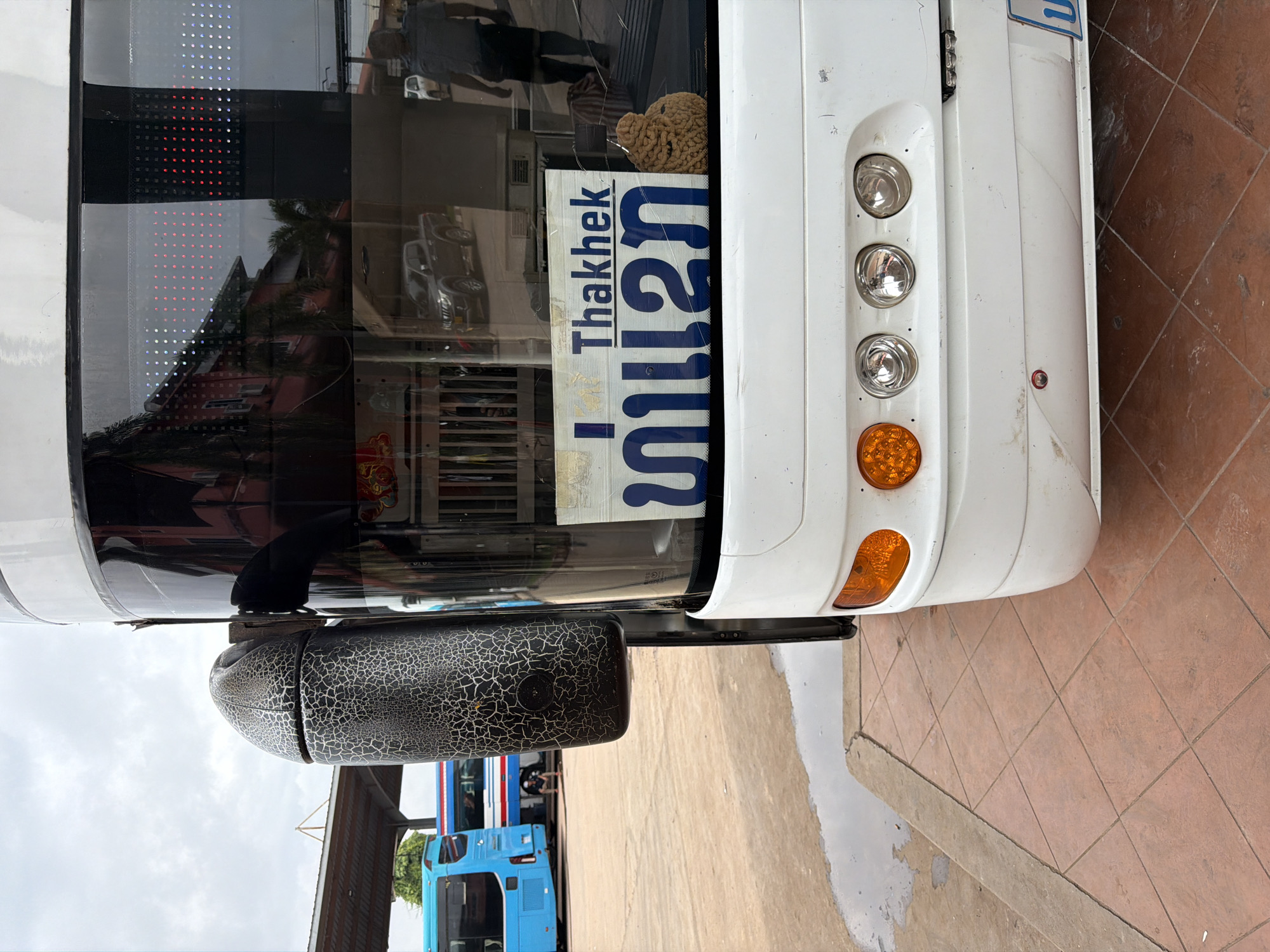 A close-up, low-angle shot shows the rear of a white tour bus parked outdoors at a bus station in Phakhao, Laos. The bus has orange turn signals and multiple clear circular lights on its rear bumper. A sign with blue text, reading "Thakhek" in English and Lao script, is affixed to the back window. To the right of the sign is a brown, textured object, possibly a decorative item or a piece of luggage. The bus's rear view mirror, with a black, cracked, textured surface, is visible on the left side of the frame. In the background, a blue and a red-and-white bus are parked under a covered area. The ground is paved with reddish-brown square tiles, with a wet patch visible on the concrete edge to the left. The sky is overcast.