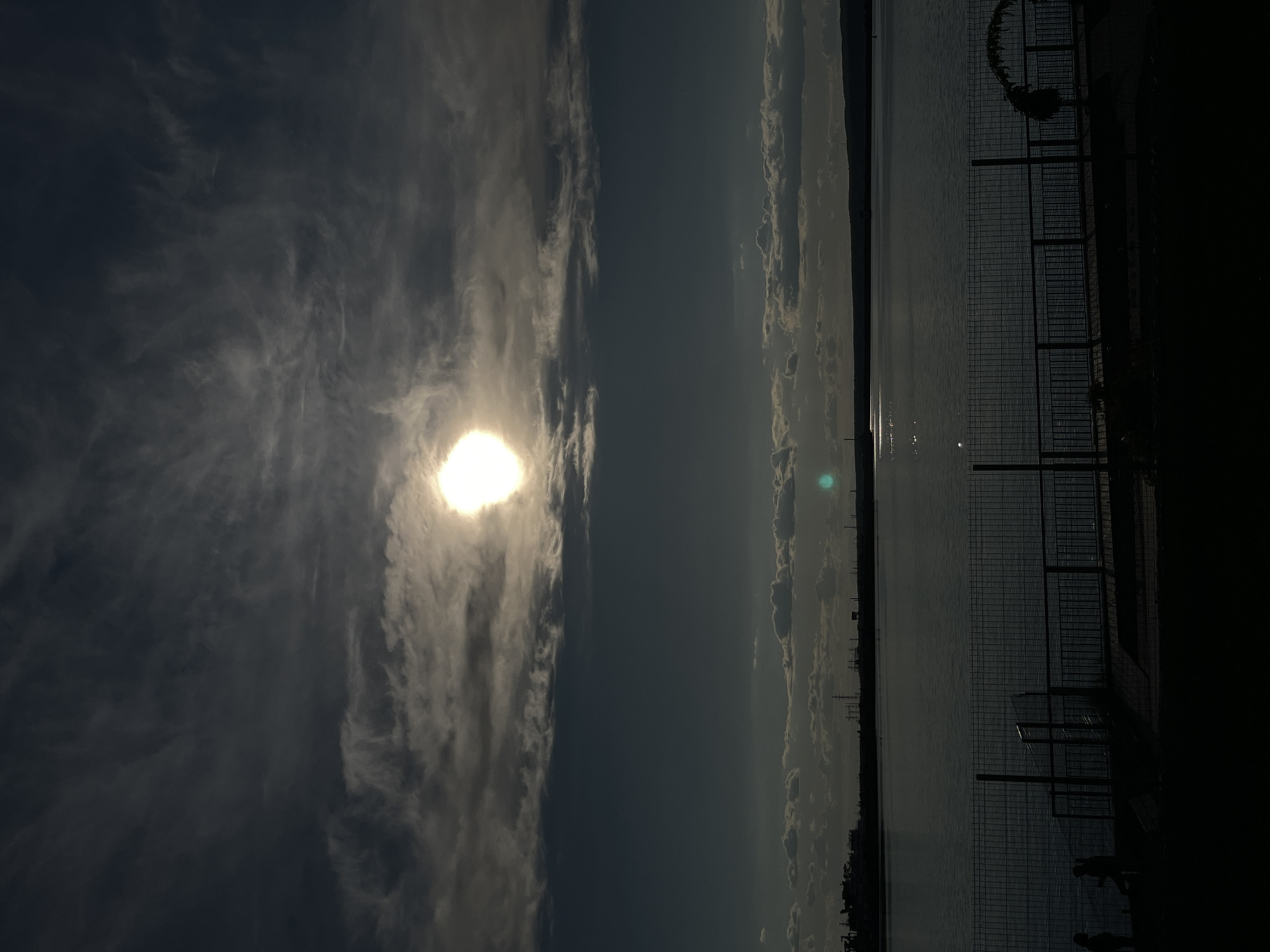 The image, when correctly oriented, presents a tranquil waterfront scene during what appears to be late afternoon or early evening, characterized by strong backlighting. The left side of the frame is dominated by a bright, intense sun, partially veiled by translucent, wispy clouds that create a soft glow. The sky around the sun is a lighter shade, transitioning to a deeper blue-grey towards the top and center, with scattered clouds visible across the horizon on the right.
A wide expanse of water, likely a lake or sea, occupies the right half of the image, reflecting the distant light in shimmering patches. Along the far horizon, a faint landmass with some indistinct structures, possibly buildings or power lines, can be seen. The immediate foreground on the bottom right features the silhouette of a shoreline area, including a prominent metal mesh fence running horizontally. Behind this fence, two small, dark figures, likely people, are visible, standing by the water's edge. Other dark, vertical structures, possibly railings or posts, are also silhouetted against the water and sky. The strong backlighting from the sun creates dramatic shadows in the foreground, enhancing the depth of the scene. No discernible text is visible in the media. This peaceful waterfront setting is located in Chaikovsky, Russia.