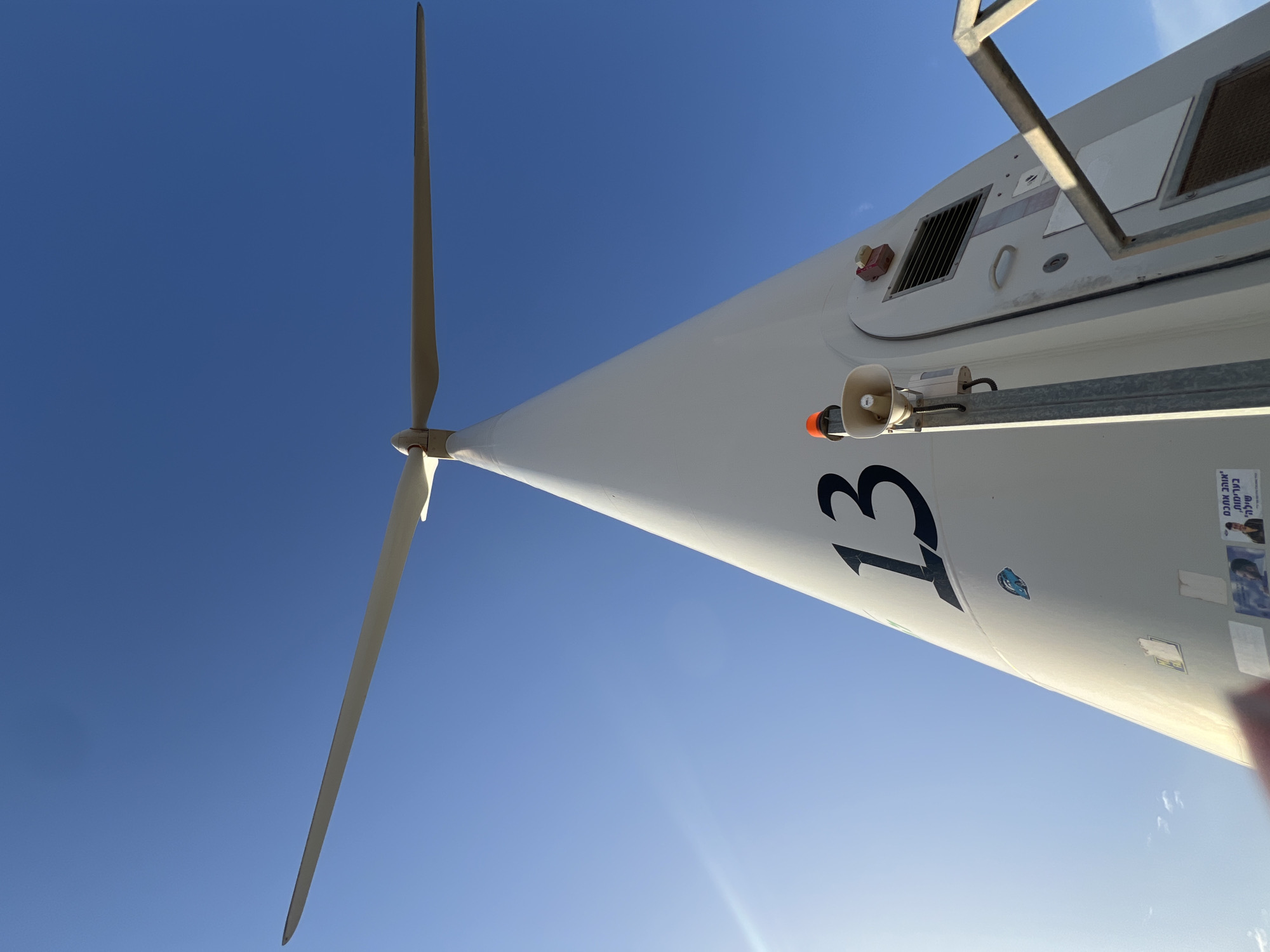 A low-angle shot of a wind turbine against a clear blue sky. The turbine, marked with the number "13", appears to be located in Emek HaMaayanot Regional Council, Israel. The blades are visible, and a speaker is mounted on the turbine structure. Some stickers and labels are on the side of the turbine.
