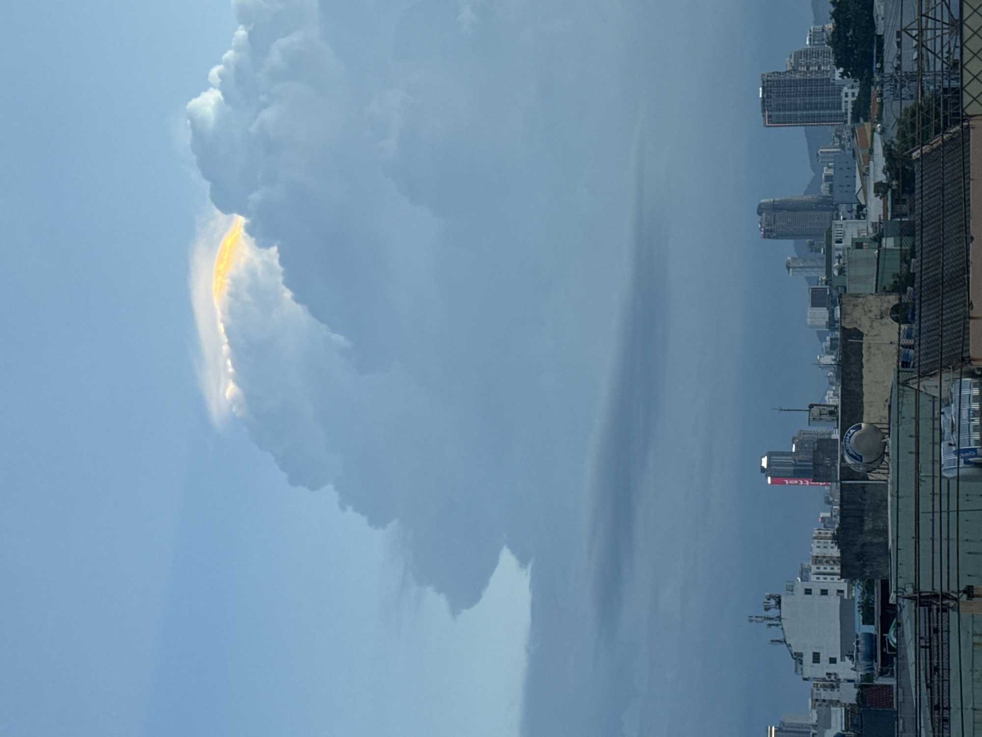 In Đà Nẵng, Vietnam, a city skyline is visible under a dramatic sky. Towering clouds dominate the upper portion of the frame, with a vibrant, golden-yellow light illuminating a section of the cloud's edge, suggesting a sunset or sunrise phenomenon. Below the clouds, a dense cityscape unfolds, characterized by various buildings of different heights and architectural styles. Prominent structures include modern high-rises with illuminated windows, and lower-rise buildings that appear to be residential or commercial. Several buildings are topped with water tanks, a common sight in urban Vietnamese areas. The scene captures the interplay of natural light and the urban environment, creating a visually striking contrast between the celestial display and the man-made structures. The image does not depict any individuals.
