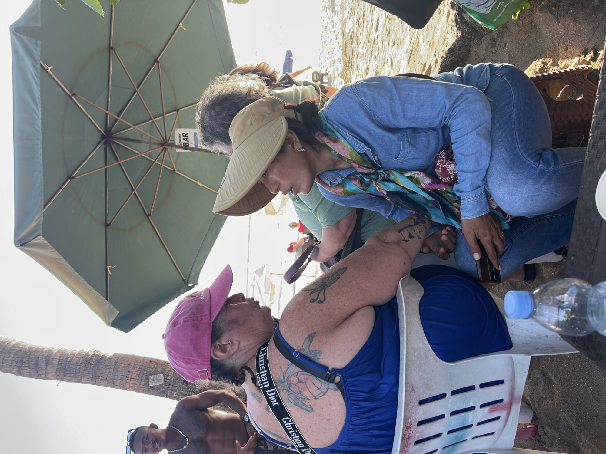In Kuta, Indonesia, at a beachside establishment, two women are engaged in conversation while seated at a table under a large green umbrella. The woman on the left, wearing a pink baseball cap and a blue dress, has her back to the camera. Her upper body is adorned with several tattoos, including a prominent sea turtle on her back. A black strap with "Christian Dior PARIS" printed in white runs across her shoulder. The woman on the right, facing the camera and wearing a tan sunhat and a denim shirt, has a colorful scarf tied around her neck. She is holding a smartphone in her hand. Both women are seated on chairs, with a plastic bottle of water visible on the table in the foreground. The background shows a sandy beach with the ocean and a palm tree, indicating an outdoor setting. A shirtless man with a chain necklace is visible to the left, standing near the palm tree.