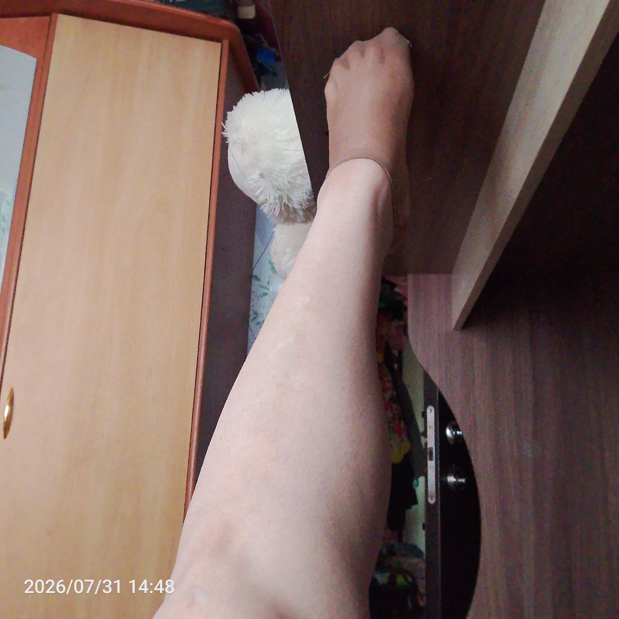 A person's leg, covered by sheer hosiery, is resting on a dark wooden surface. A portion of a white stuffed animal is visible behind the leg. To the left, a light brown wooden cabinet with a gold handle is present. The bottom left corner of the image displays a timestamp: "2026/07/31 14:48". The environment appears to be indoors, possibly a room with furniture.