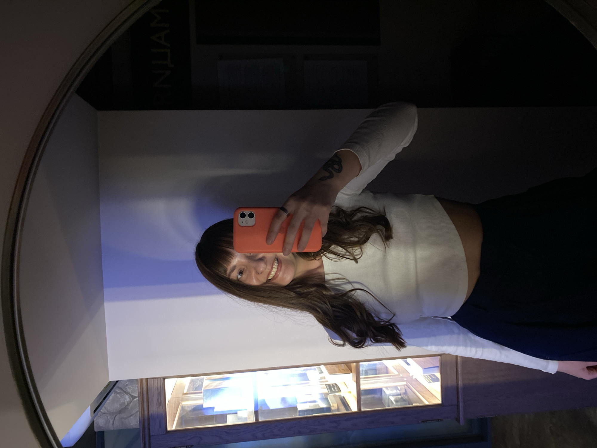A woman is reflected in a round mirror, holding a smartphone in front of her face to take a picture. She is smiling. She is wearing a white, long-sleeved crop top and dark-colored pants. The reflection includes a display case with illuminated objects inside, and a wall with the letters "НЦИАМ" visible. The location is likely the Museum of the History of the Worker's Movement in Votkinsk, Russia, as the abbreviation "НЦИАМ" potentially stands for "Научно-культурный и информационный центр Архивное дело Музейное дело" (Scientific, cultural and informational center archival affairs museum business) which could be the full name of the museum.