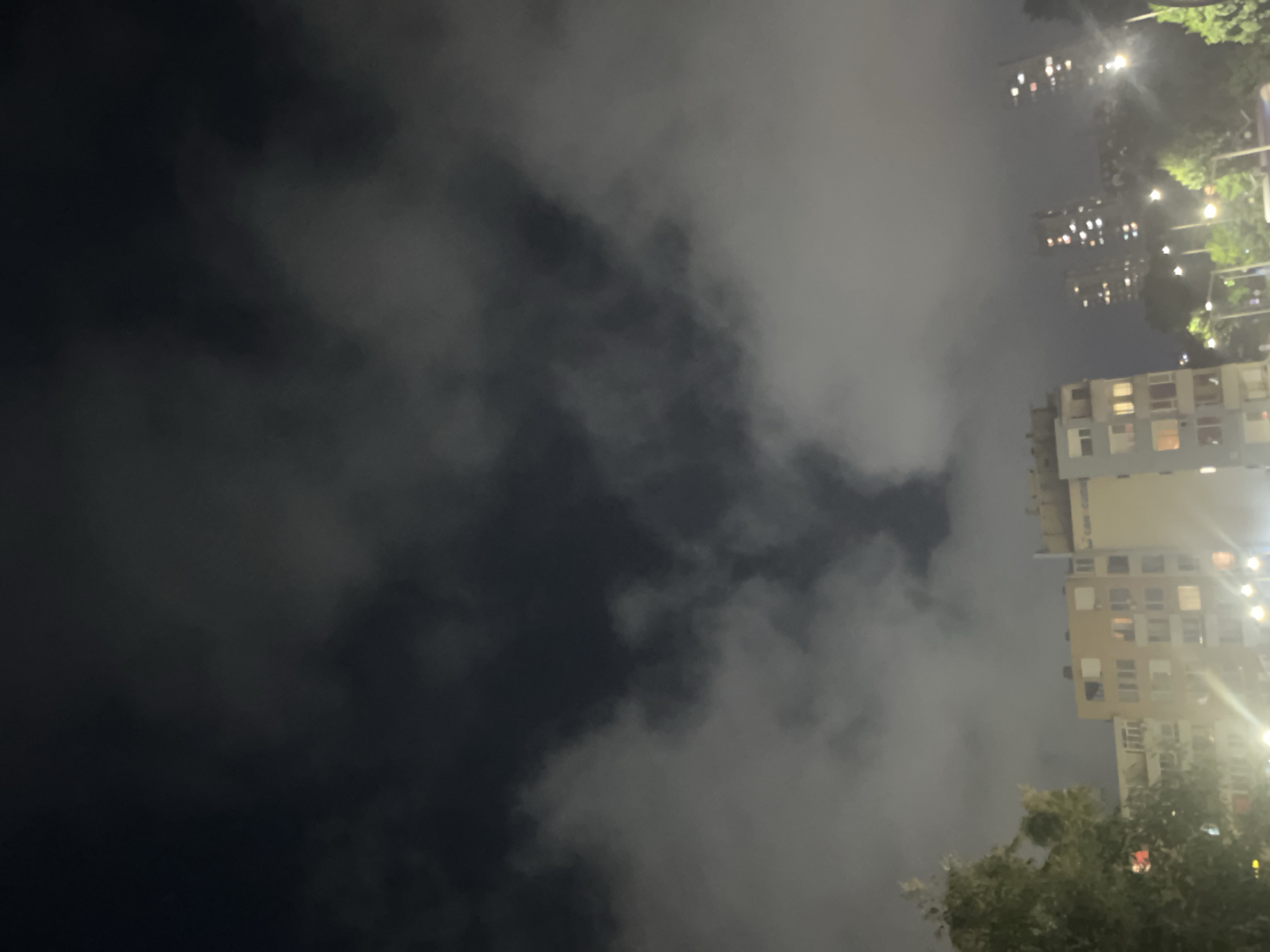 The image depicts a nighttime urban scene in Burgas, Bulgaria, characterized by heavy, low-lying cloud cover and artificial illumination. The sky is predominantly obscured by thick, dark grey clouds, but a distinct, roughly V-shaped or heart-shaped void of deeper black sky is visible near the upper center, suggesting an opening in the cloud mass. Below the clouds, an array of artificial lights from buildings and streetlamps brightens the lower half of the frame.
In the mid-ground, a multi-story apartment building with a light beige or yellowish facade and white balconies occupies the left and central portion. Numerous windows, some illuminated with warm light, are visible across its structure. To the immediate left of this building, a large, leafy tree is partially visible. Further in the background on the right, several taller, more distant buildings, likely residential towers, rise into the hazy night, their numerous windows glowing with light. The foreground and mid-ground also show several streetlights emitting a warm, yellowish glow, illuminating portions of the ground and foliage. Trees and shrubbery are scattered across the lower right, partially obscuring the view of the distant buildings. No individuals or specific actions are discernible.