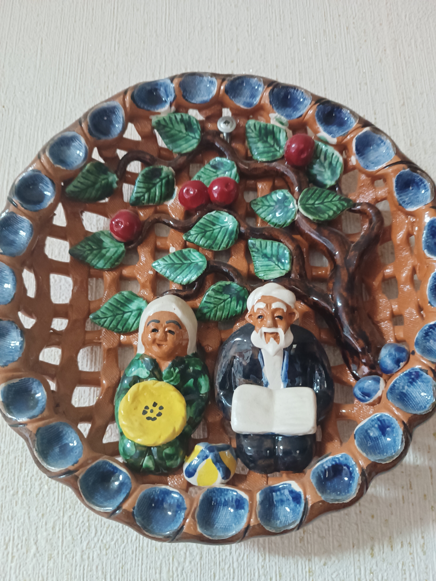 The image shows a decorative ceramic plate depicting an elderly couple under a tree bearing red fruit. The plate has a brown latticework background with circular blue details around the rim. The couple appears to be of Central Asian or Middle Eastern descent. The woman is on the left, wearing a green patterned garment and holding a yellow object with black markings. The man is on the right, wearing a dark suit and holding a white rectangular object, possibly a book. A yellow and blue object sits between them. The plate is hanging on a wall with a textured, possibly painted, surface.
Based on visual evidence, there's no immediate connection to the city of Kineshma, Russia. More information would be necessary to make that connection.