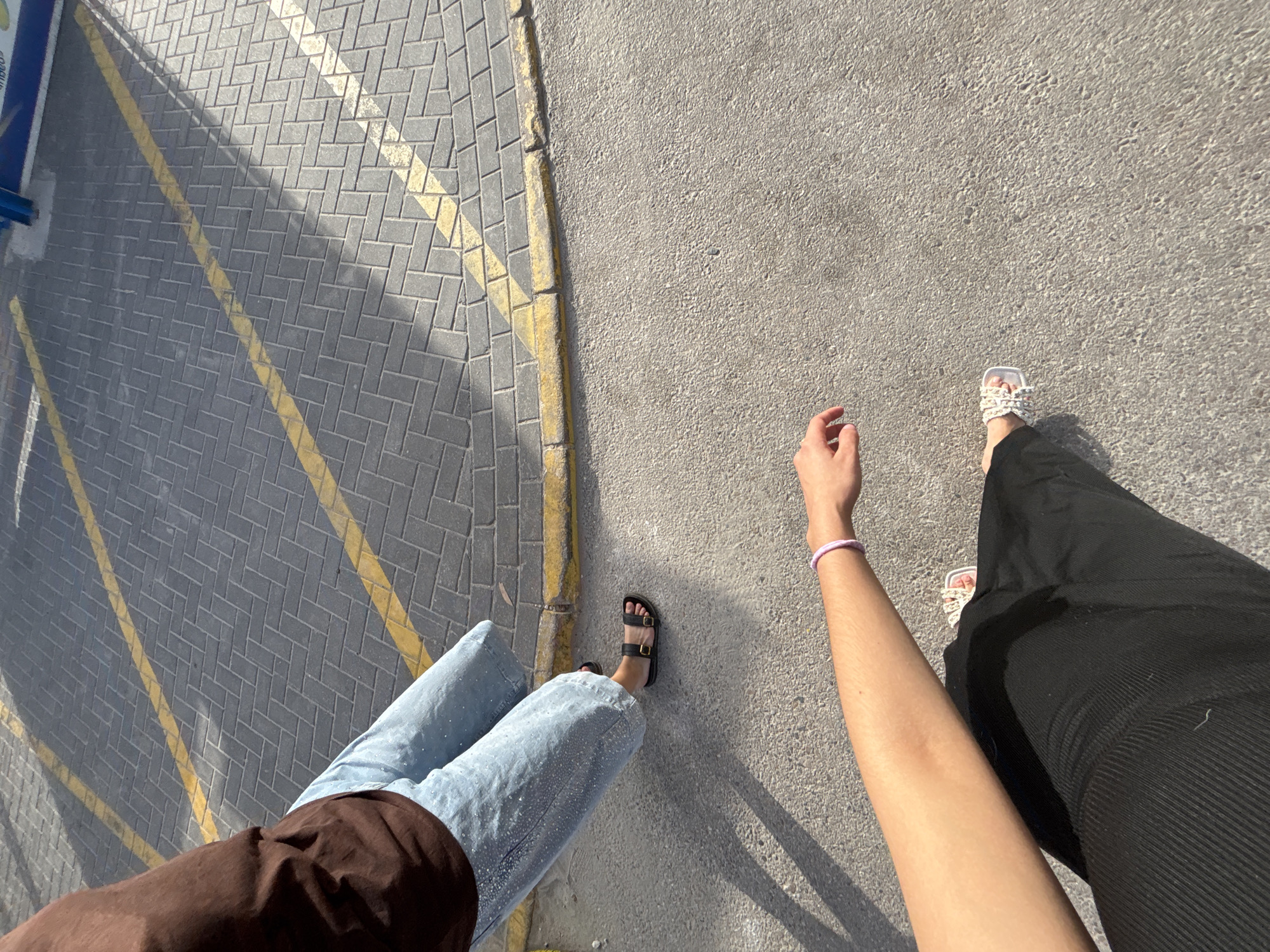 The image shows a low-angle view of two people's lower bodies and legs in an outdoor urban setting. The ground consists of a paved area with yellow and white painted lines, suggesting a parking lot or walkway. One person is wearing light blue, wide-leg jeans with a sparkling texture and black flat sandals. This individual is standing near a curb with the paved area of interlocking grey bricks behind them. The second person, closer to the camera and on the right side of the frame, is wearing a black garment that appears to be a skirt or wide-leg pants, and white sandals with a woven texture. Their left arm, with a pink bracelet, extends into the frame from the bottom left corner. The scene is lit by direct sunlight, casting distinct shadows on the ground. The location is identified as Urbanización Cabo Roig, Spain.