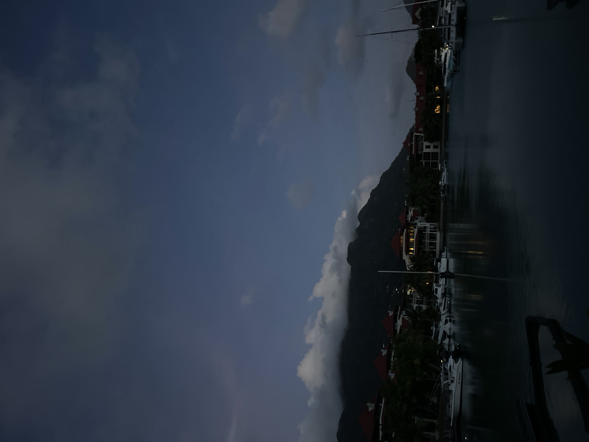 The image depicts a twilight scene in Providence, Seychelles. The foreground is dominated by a dark body of water reflecting the dim light. In the middle ground, a line of boats is moored along a waterfront. These appear to be small to medium-sized recreational boats and yachts. Behind the boats, several buildings with red-tiled roofs and illuminated windows are visible, suggesting a residential or resort area. Lush green palm trees are interspersed among the buildings. The background features a dark, mountainous landscape partially obscured by heavy, low-lying clouds. The sky above is a deep blue, transitioning to a lighter shade near the horizon, with a few scattered clouds. The overall impression is one of a tranquil, early evening setting in a coastal town.