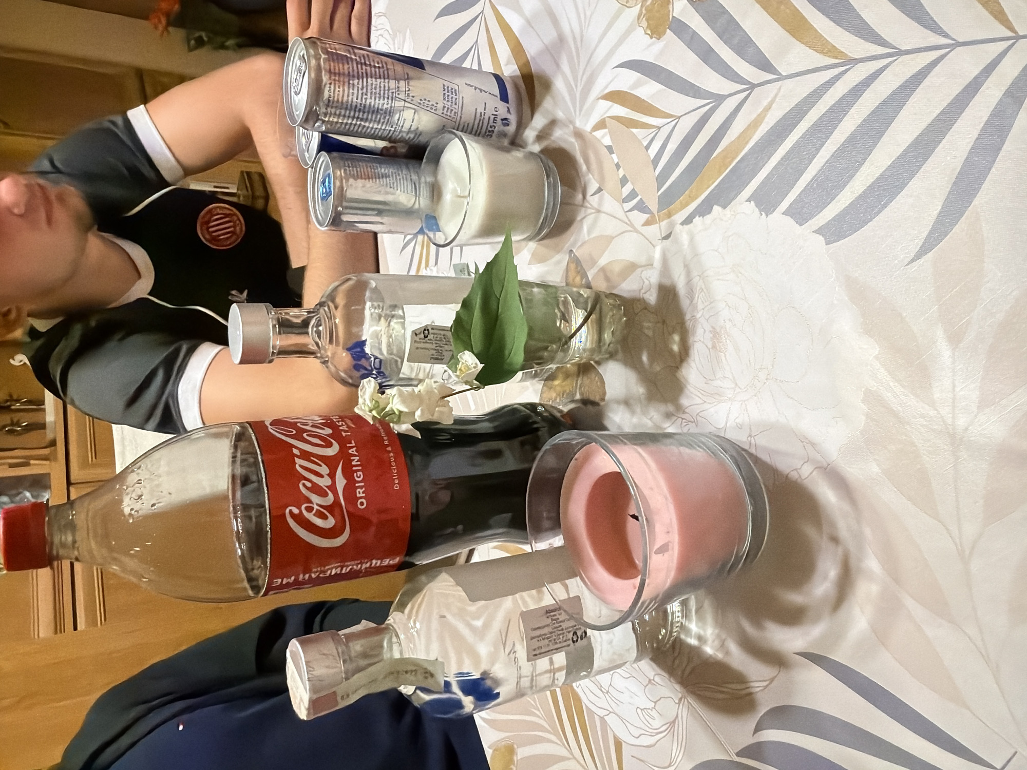 The image depicts a cluttered table scene, presumably indoors, in Burgas, Bulgaria. A fair-skinned person wearing a black and grey soccer jersey is partially visible on the left side of the frame. On the table are two metallic cans and two clear glass candle holders, one with a white candle and the other with a pink candle. There are two bottles of alcohol and a glass bottle of Coca-Cola. The table covering is a light-colored fabric with a floral and leaf pattern. A single green leaf and white flowers adorn one of the liquor bottles. A cabinet is partially visible in the background.