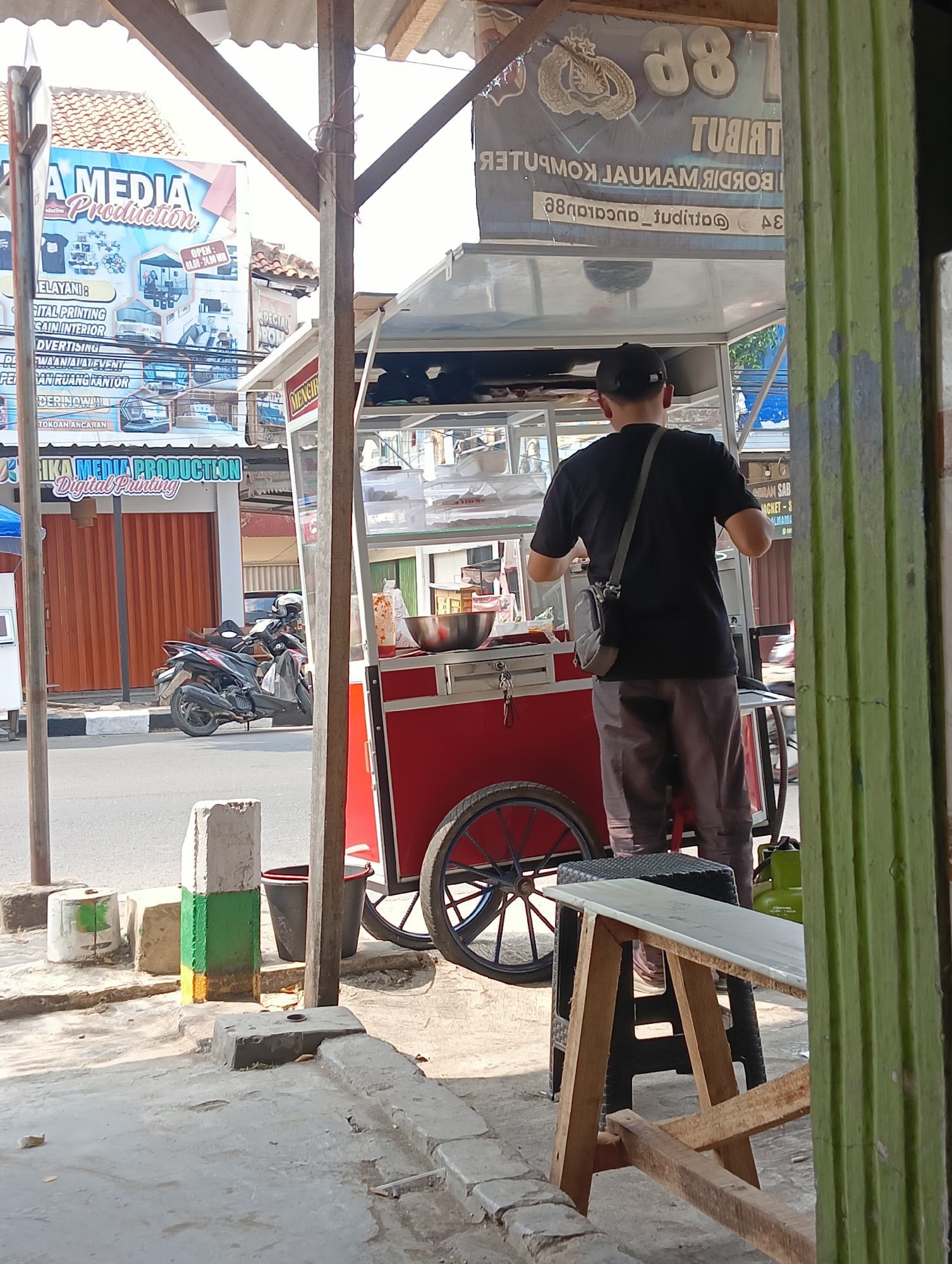 A person wearing a black t-shirt and gray pants stands behind a red food cart. The person has a black baseball cap on and a grey bag slung over their shoulder. The food cart is on a paved street in front of an orange storefront. A motorcycle is parked in front of the storefront. In the background, a sign reads "A MEDIA Production." The scene appears to be located in Manggari, Indonesia.