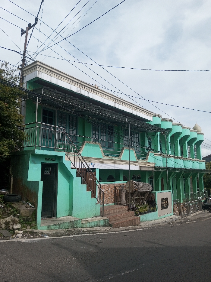 A two-story, light green building stands on the roadside. The upper level features a row of arched columns, windows with bars, and a metal-roofed balcony with a staircase. The lower level is accessed by stone steps, and a closed door is visible on the left. An old wooden drum sits near the main entrance. Overhead, a web of electrical wires crosses the sky above the building. A partial view of vegetation is seen on the left.